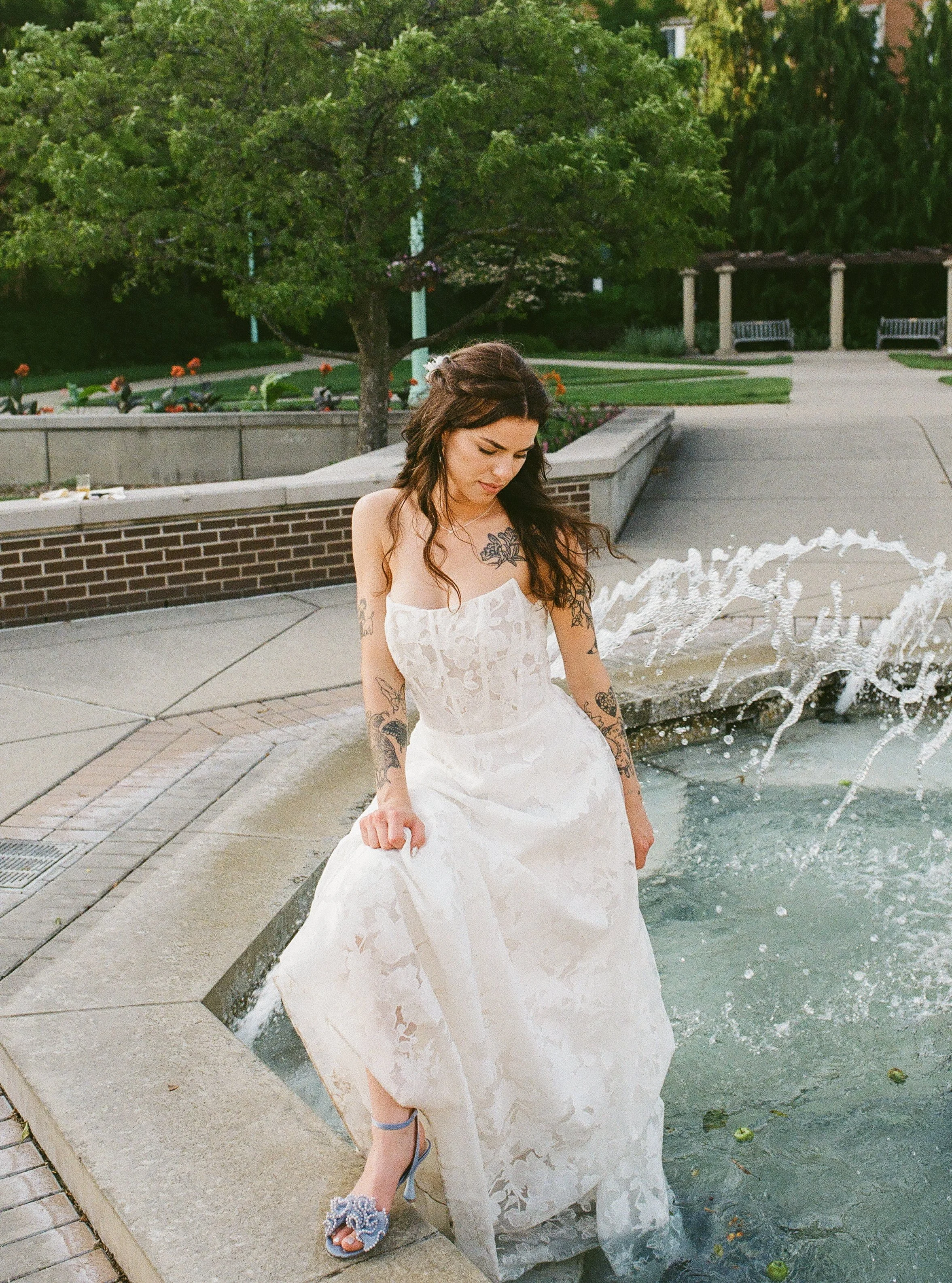 Bride standing beside a fountain holding her dress, soft sunlight highlighting the lace details in a film wedding photography moment