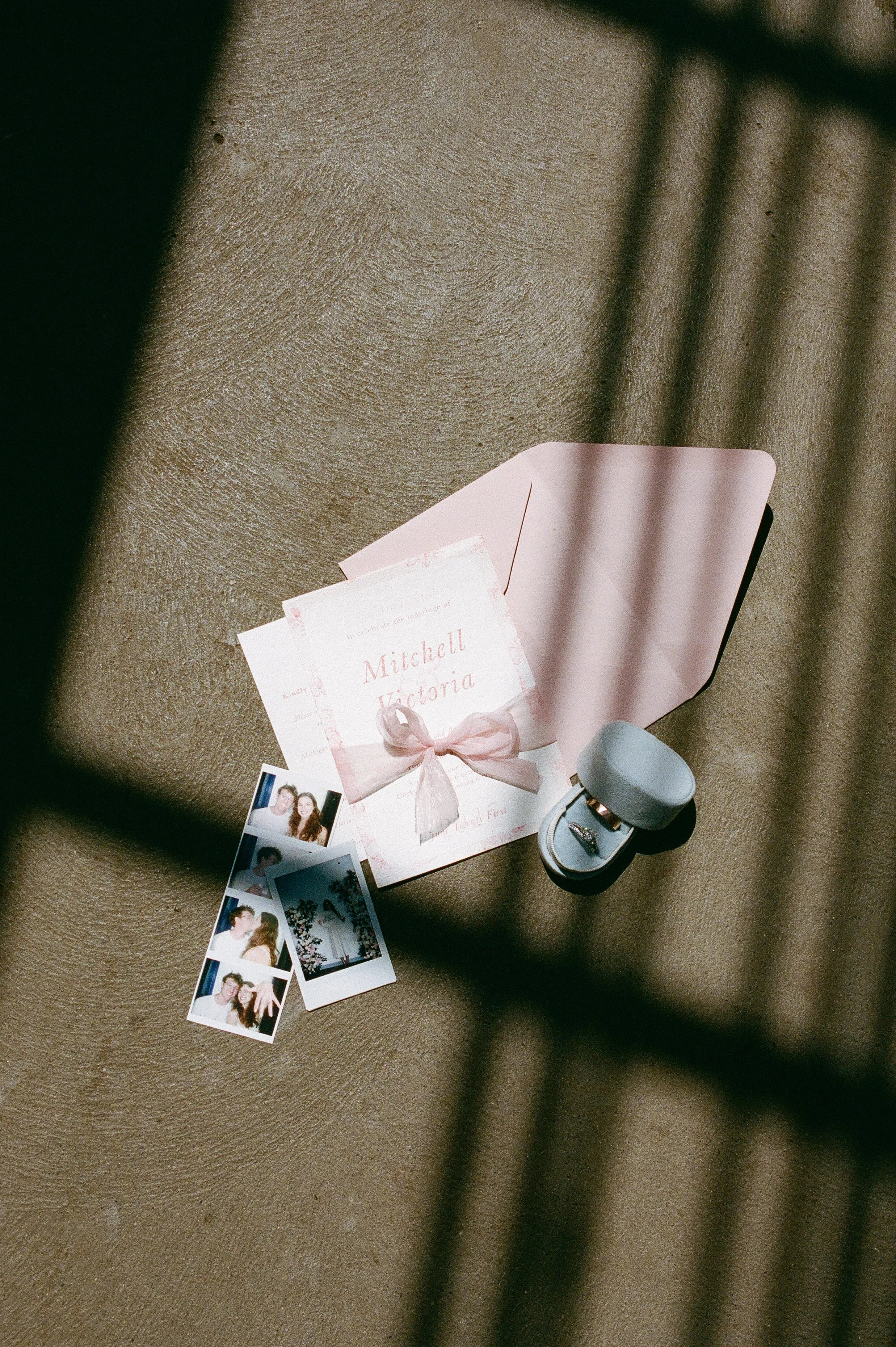 Flat lay of wedding invitation suite with ribbon, ring box, and printed photos arranged in soft natural light, styled in a film wedding photography aesthetic