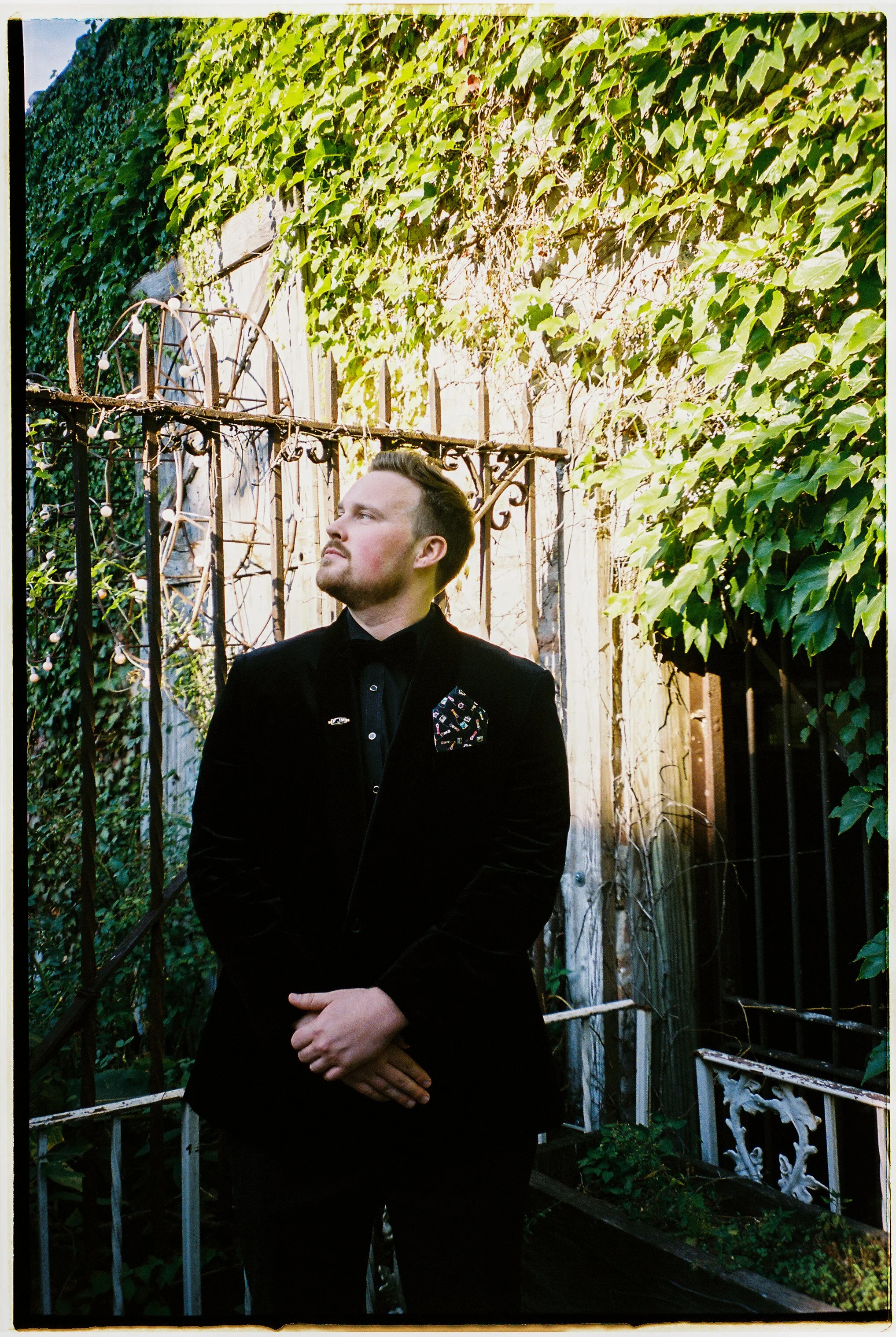 Groom portrait standing near ivy-covered walls and wrought iron gate in an outdoor courtyard
