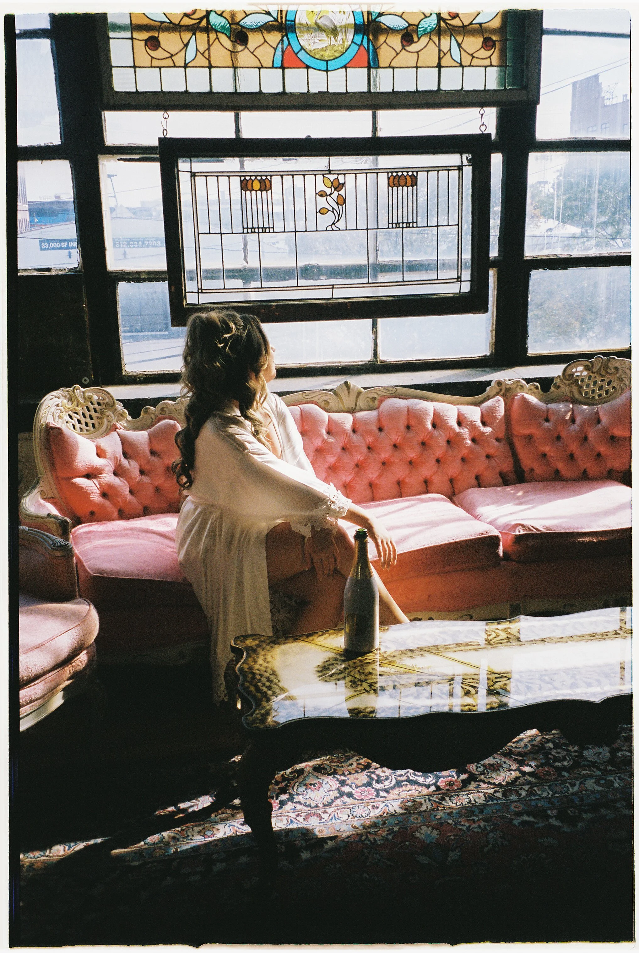 Bride in a robe sitting on a pink velvet couch by stained glass windows in warm sunlight