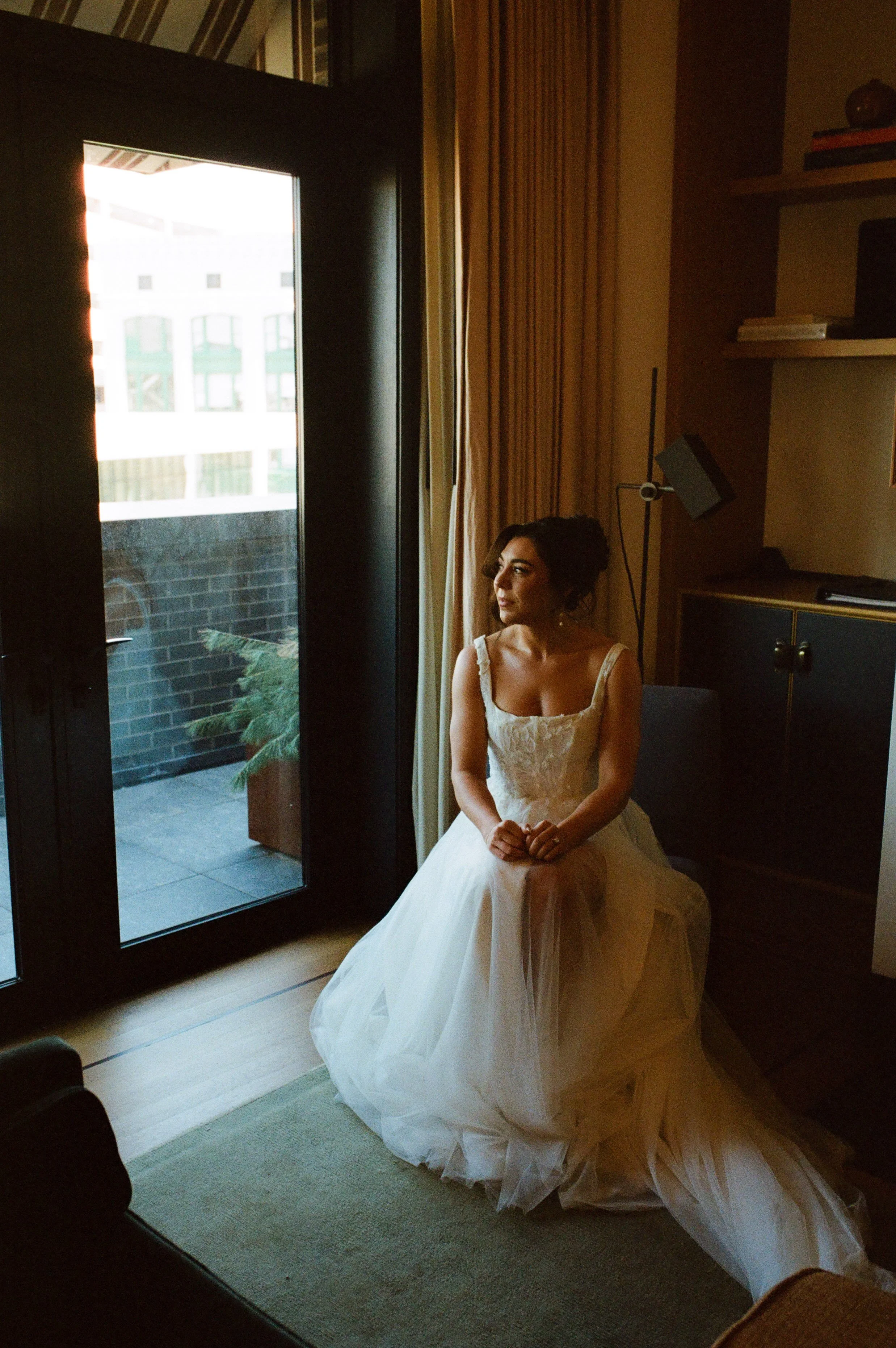 Bride sitting beside a window in a softly lit hotel suite before her wedding ceremony