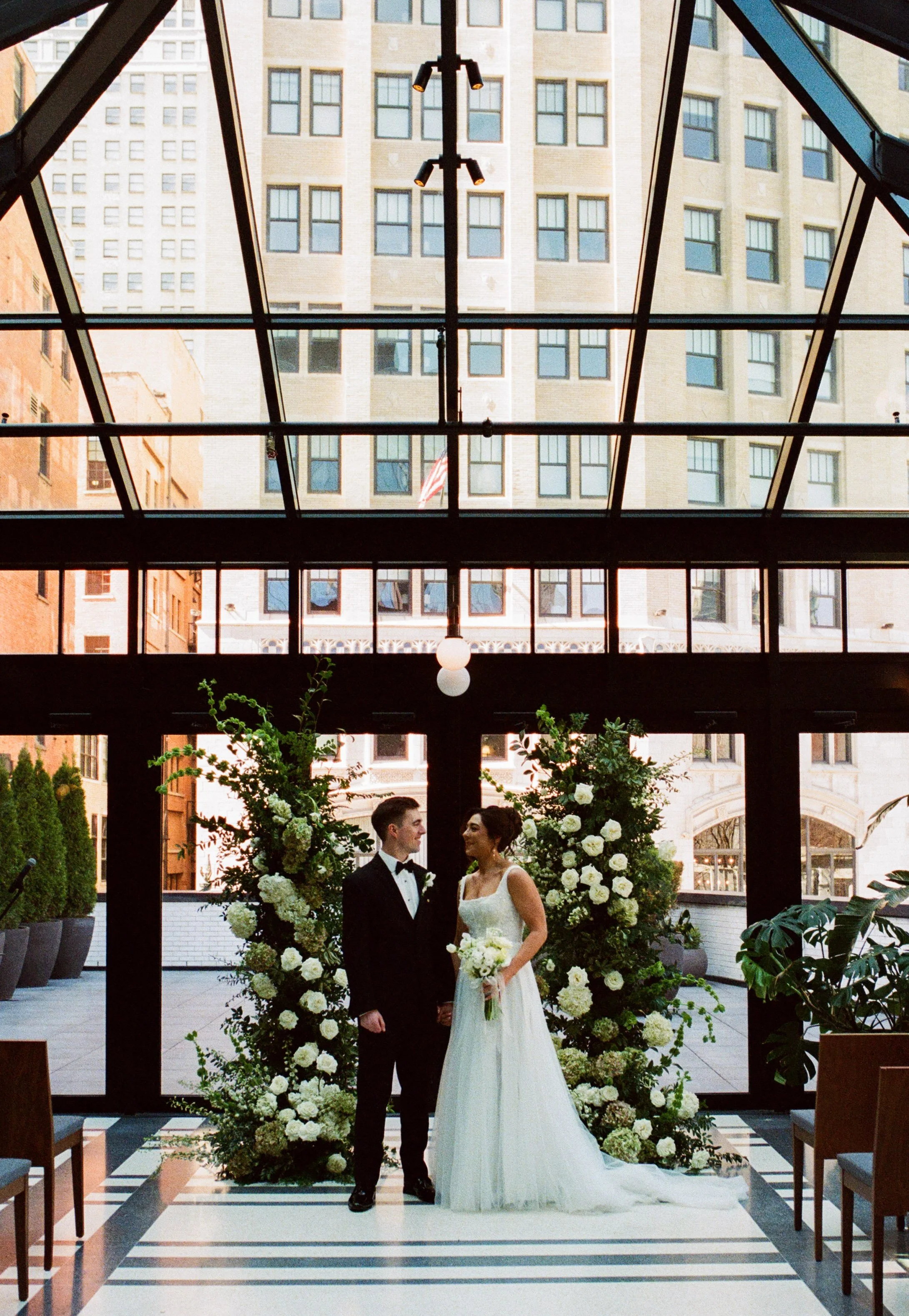 Couple standing beneath a floral ceremony installation inside Shinola Hotel with downtown Detroit views behind them