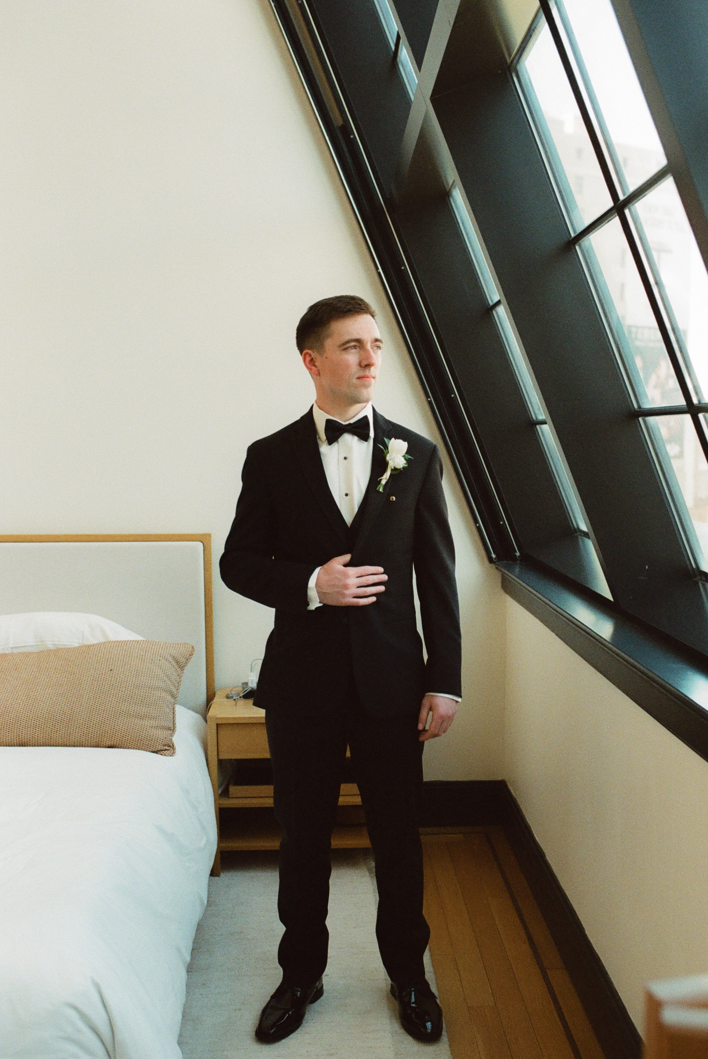 Groom in a black tuxedo standing beside large modern windows inside Shinola Hotel