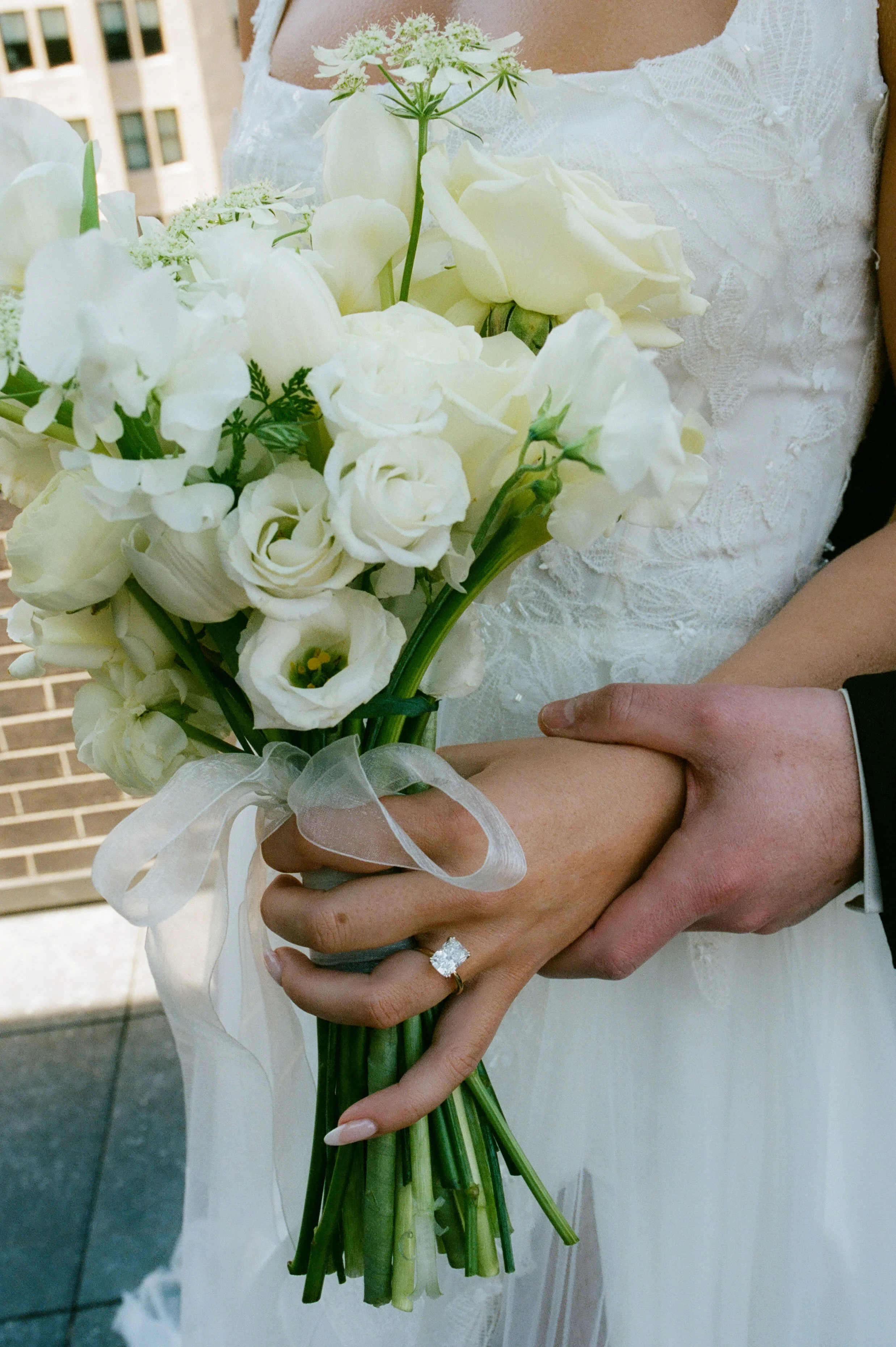 Close-up of a bride holding a white floral bouquet with her engagement ring and lace wedding dress details visible