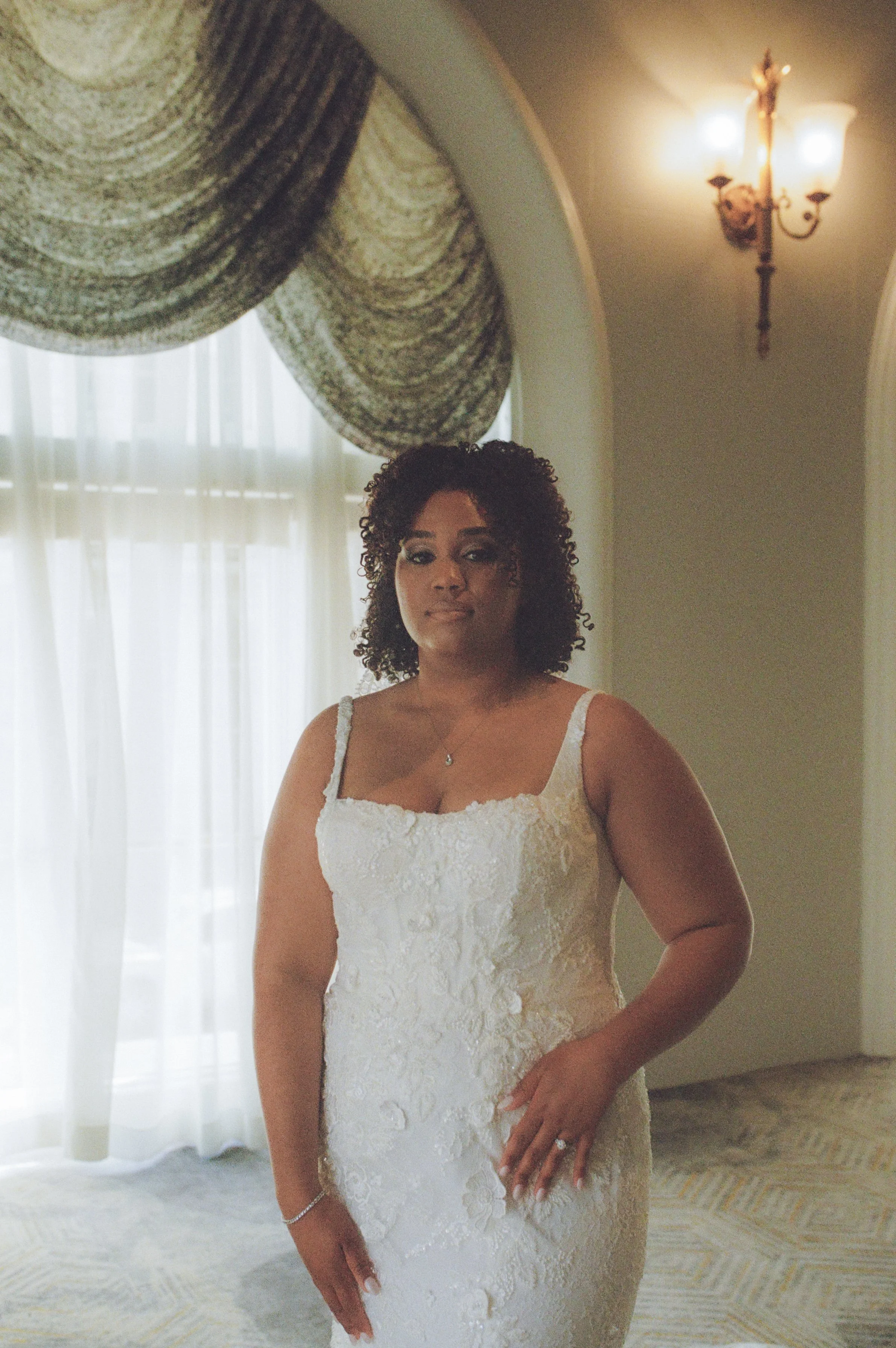 Bridal portrait of a bride standing beside tall arched windows in a softly lit ballroom captured with cinematic film photography tones