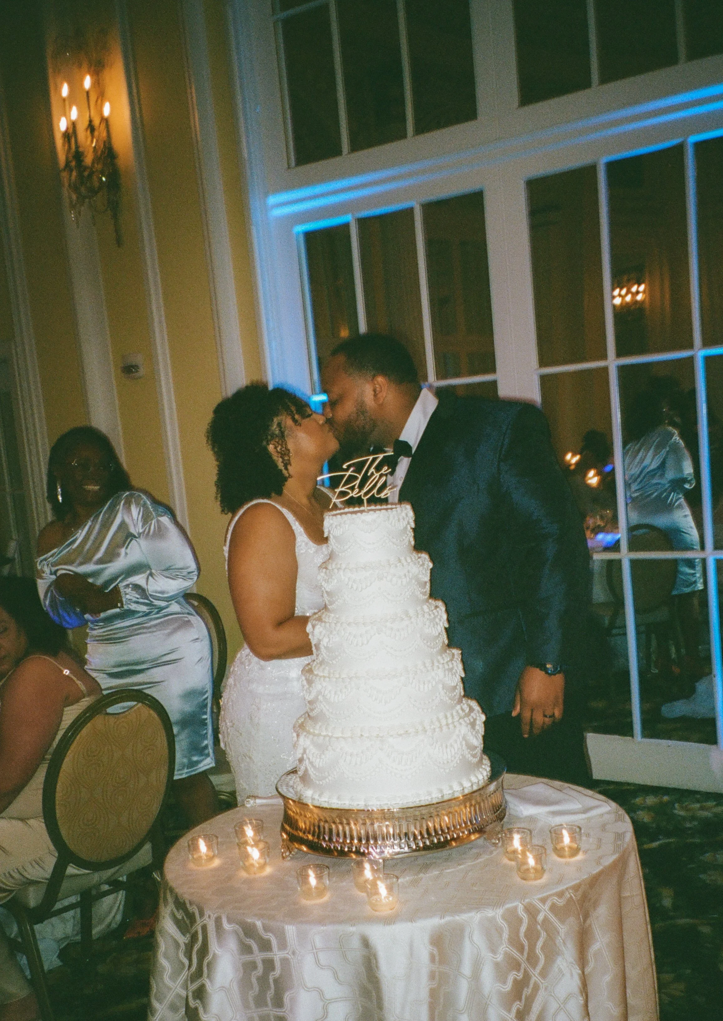 Bride and groom kissing beside a tiered wedding cake surrounded by candlelight during their reception