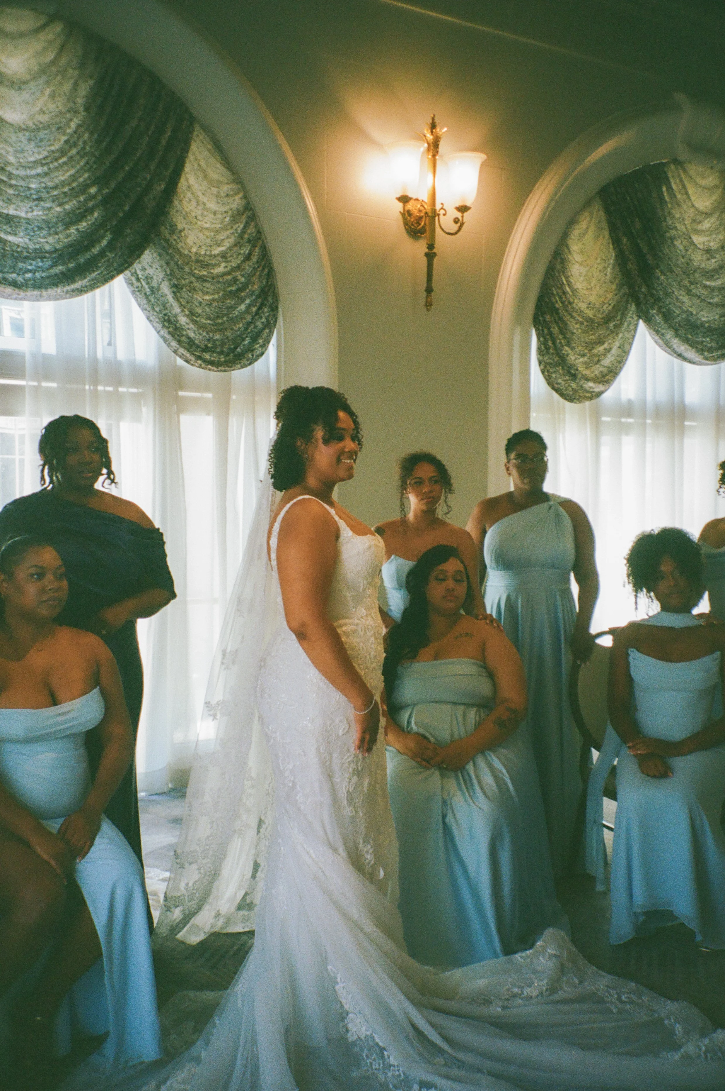 Bride posing with bridesmaids in soft blue dresses beside tall arched windows before the ceremony
