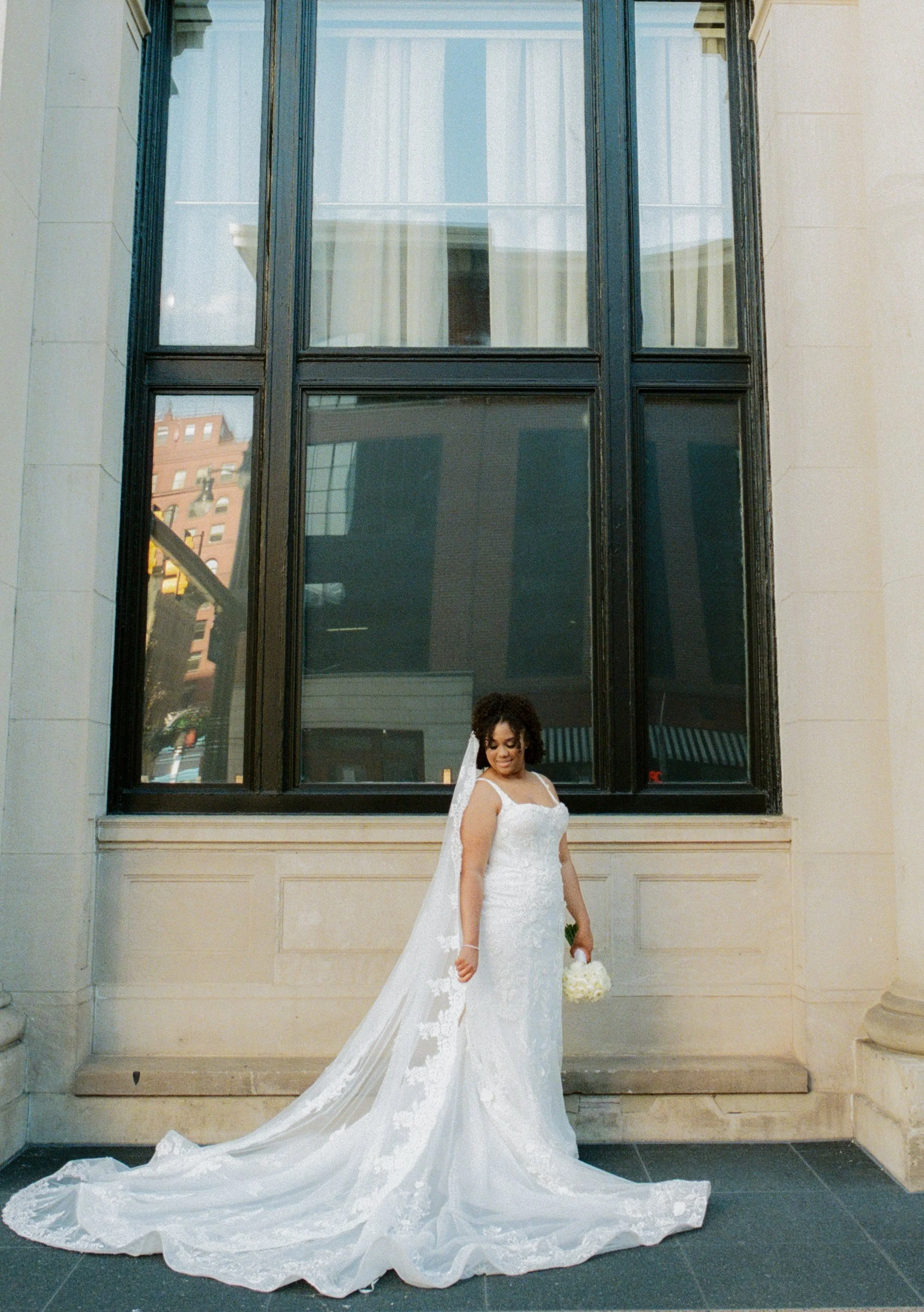 Elegant bridal portrait outside a historic hotel featuring a long lace veil and bouquet of white roses with cinematic film photography tones
