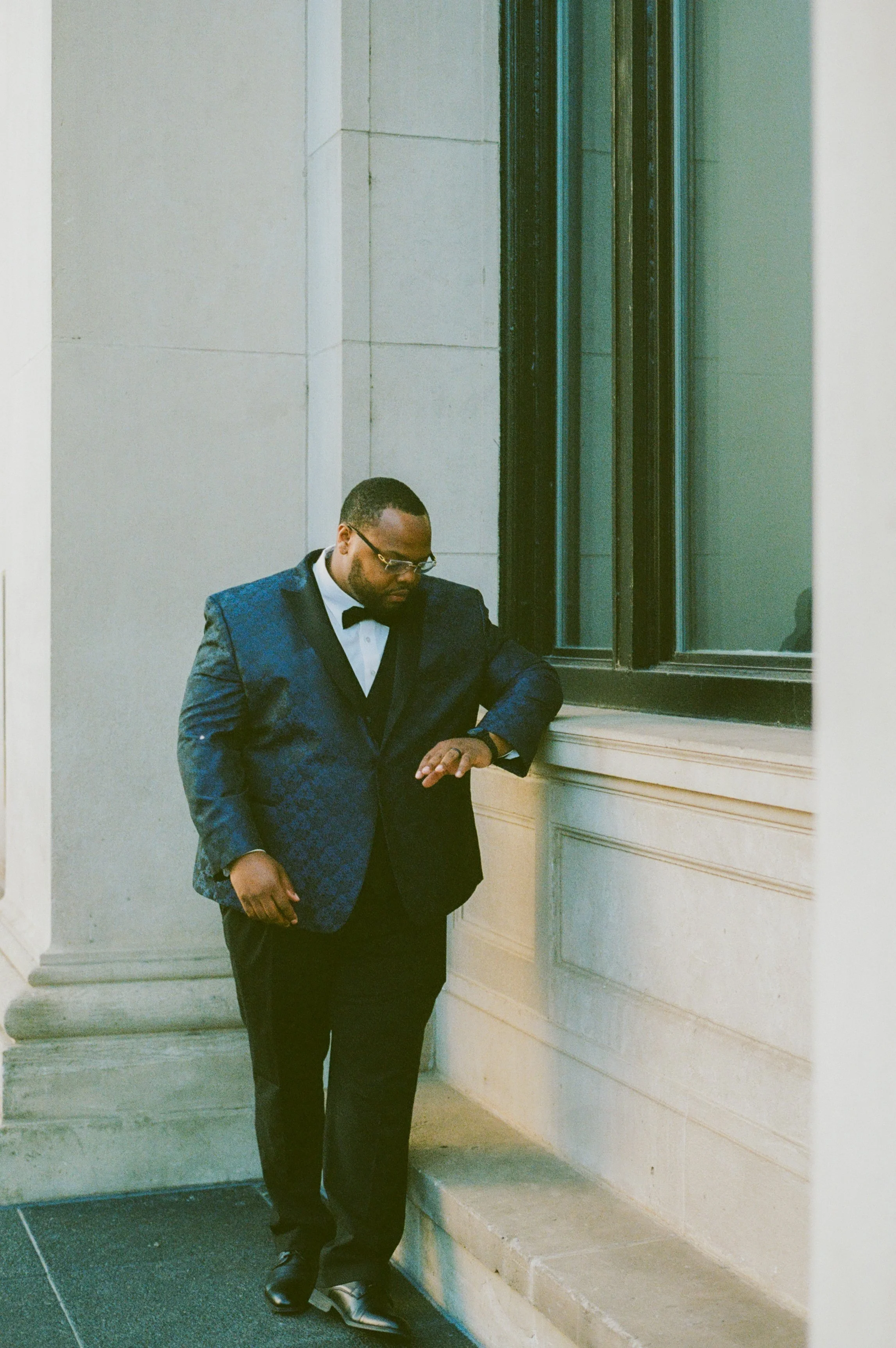 Groom portrait outside a downtown hotel wearing a navy tuxedo jacket and black bow tie captured in cinematic film photography style