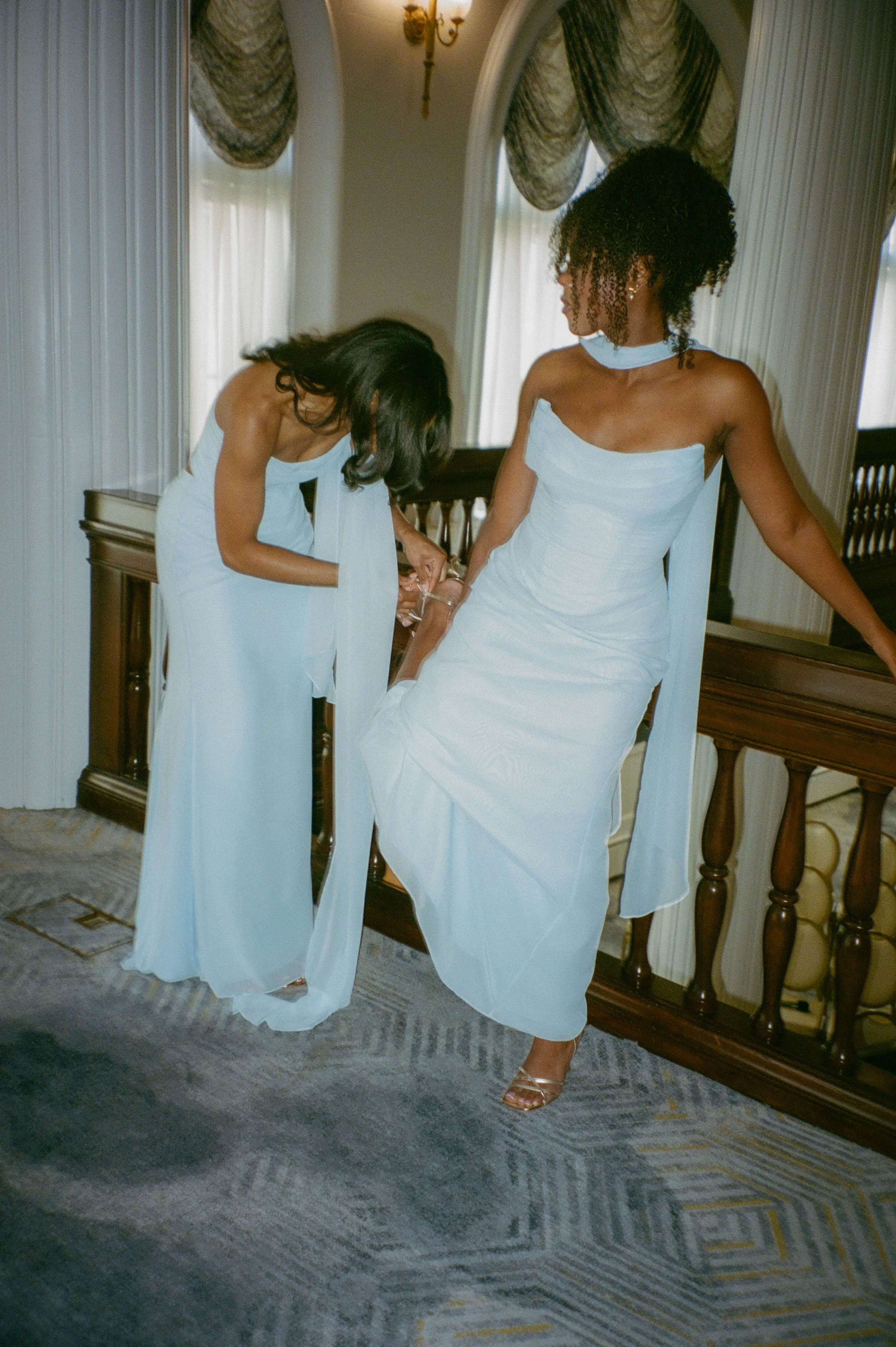 Bridesmaid helping adjust another bridesmaid’s light blue dress inside a historic hotel hallway