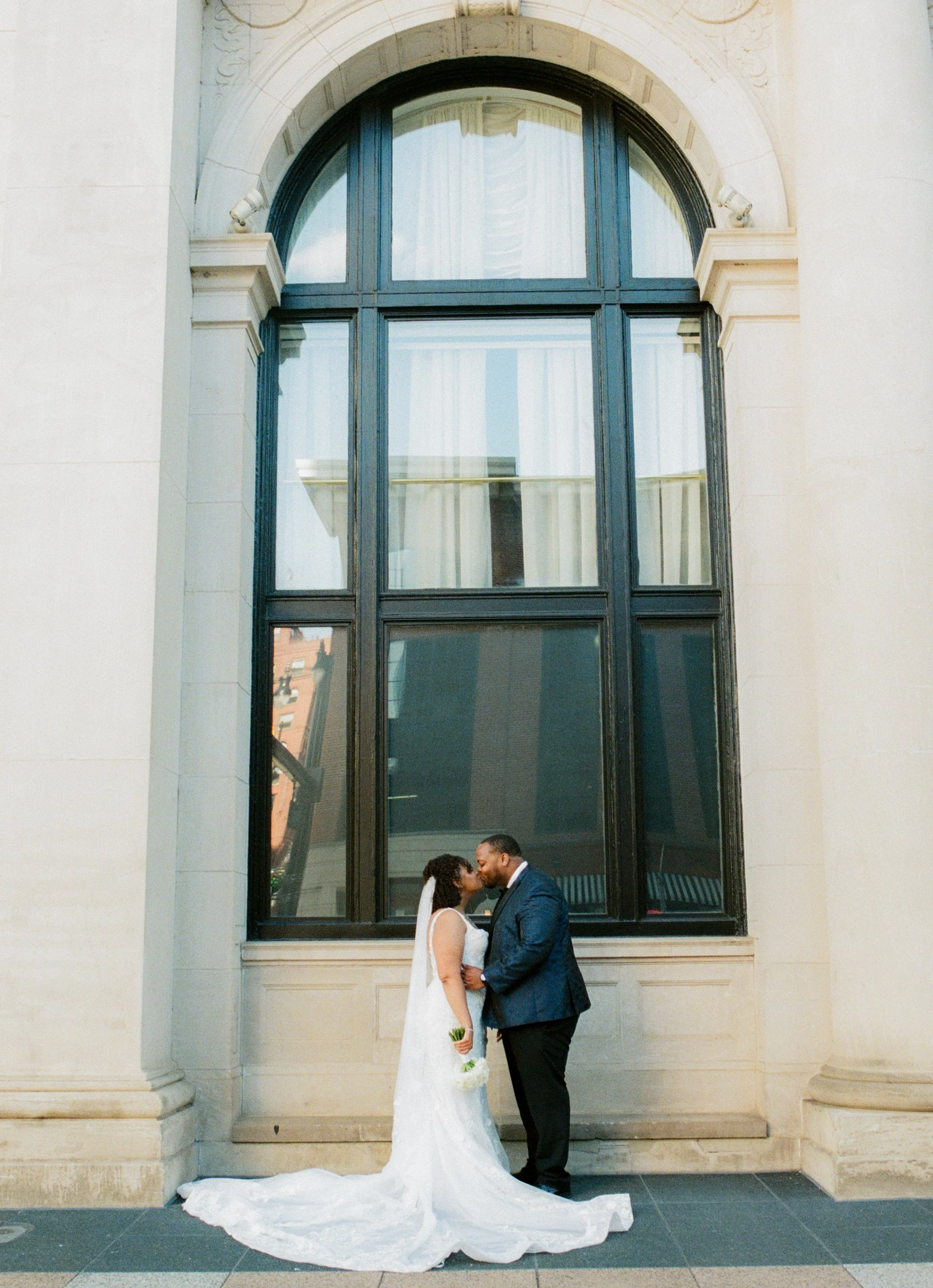 Wide wedding portrait of the bride and groom standing beneath tall arched windows outside a historic building with cinematic film photography tones
