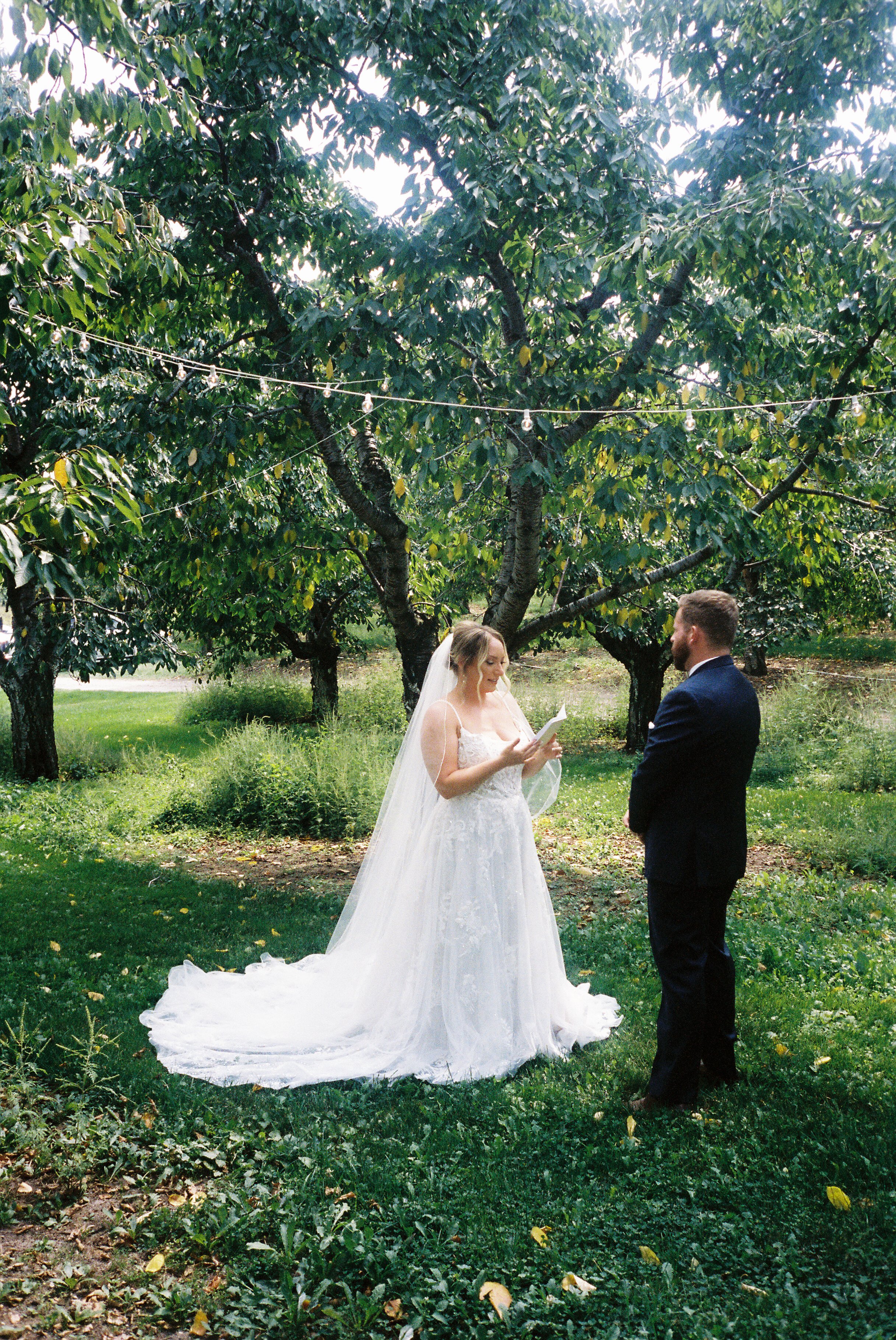 Bride reading private vows to her groom beneath mature trees before their outdoor wedding ceremony.