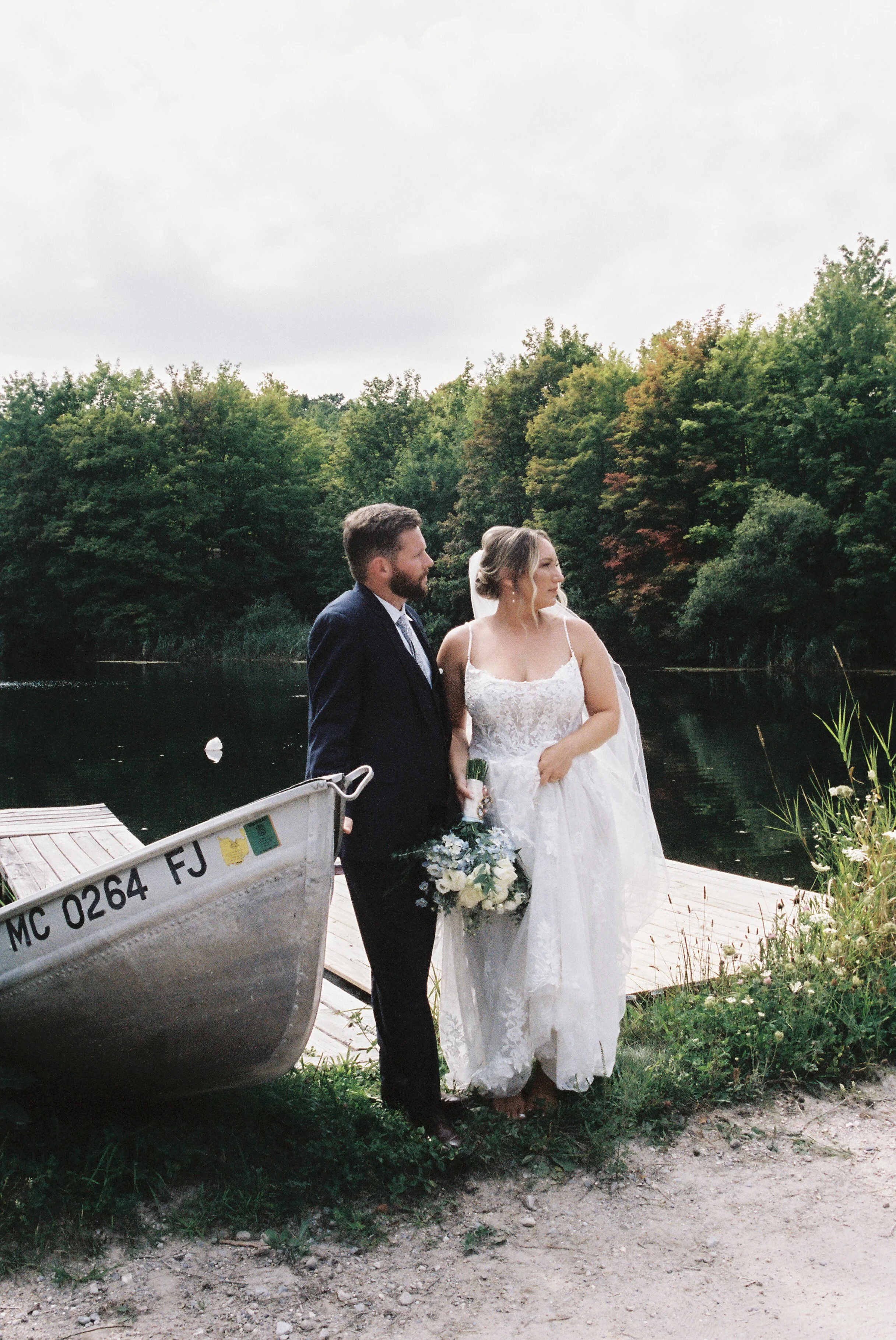 Newlywed couple standing beside a small boat and dock overlooking a peaceful lake after their wedding ceremony.
