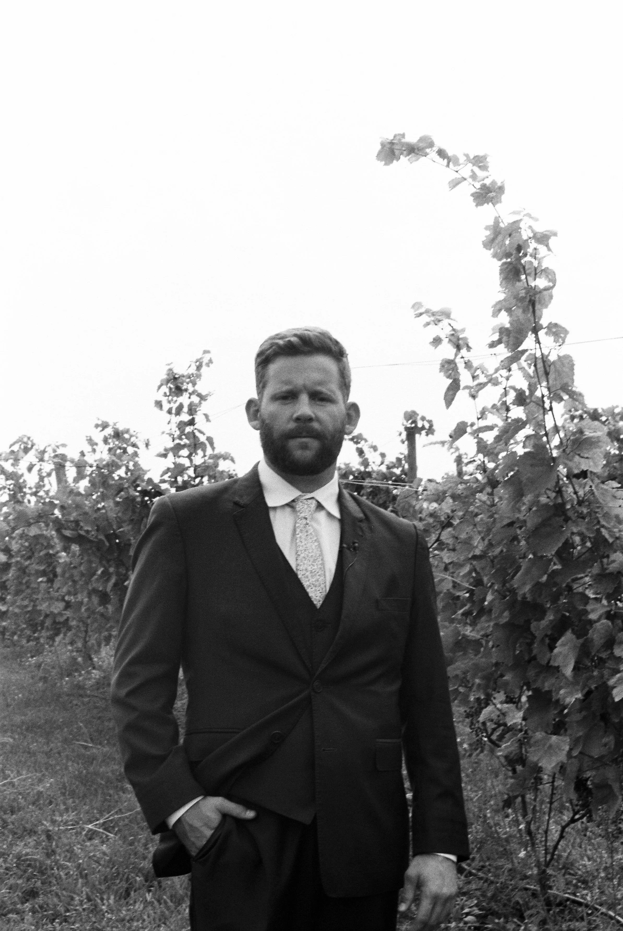 Black-and-white groom portrait standing among vineyard vines before the wedding ceremony.