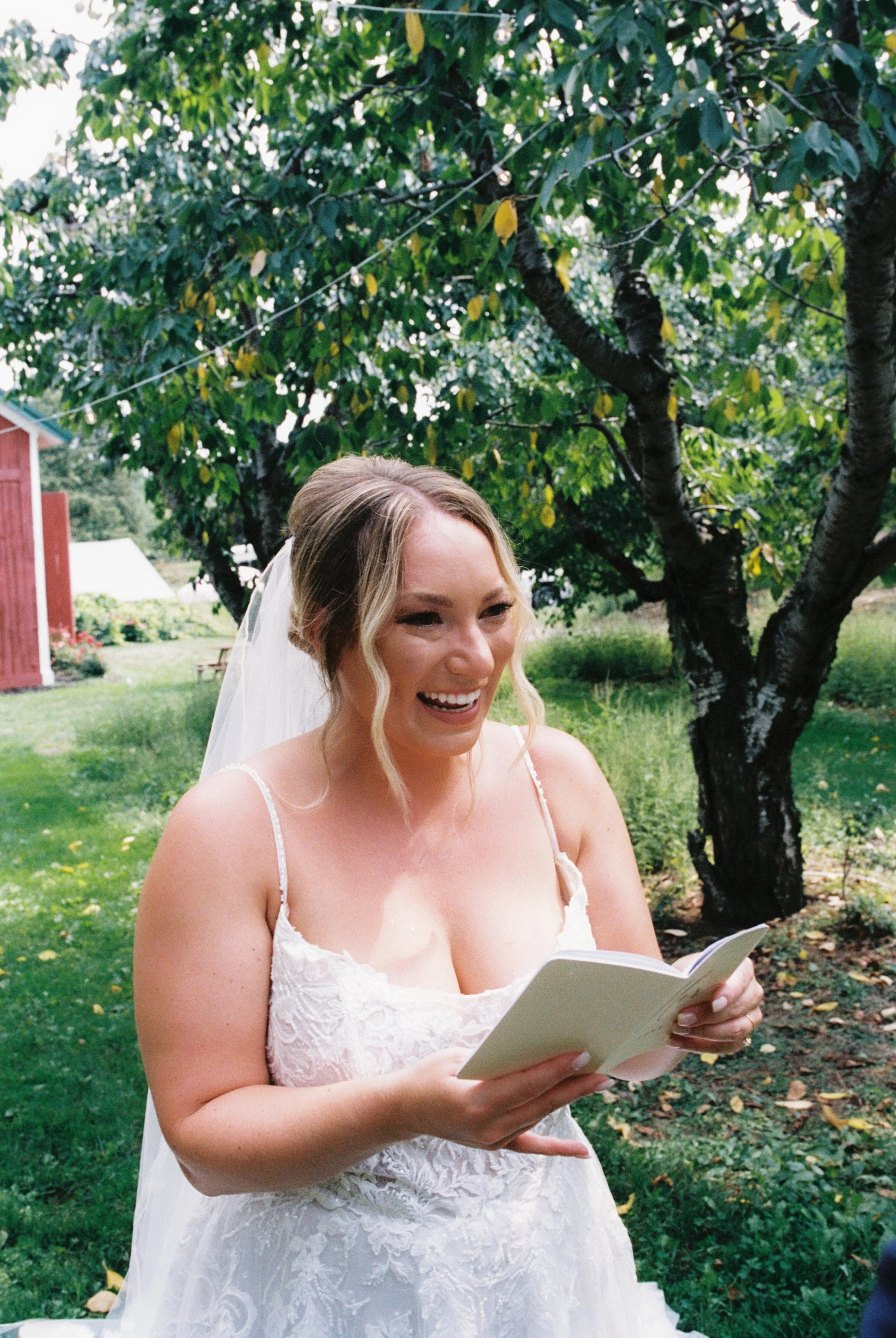 Bride smiling while reading private vows beneath orchard trees before her wedding ceremony.