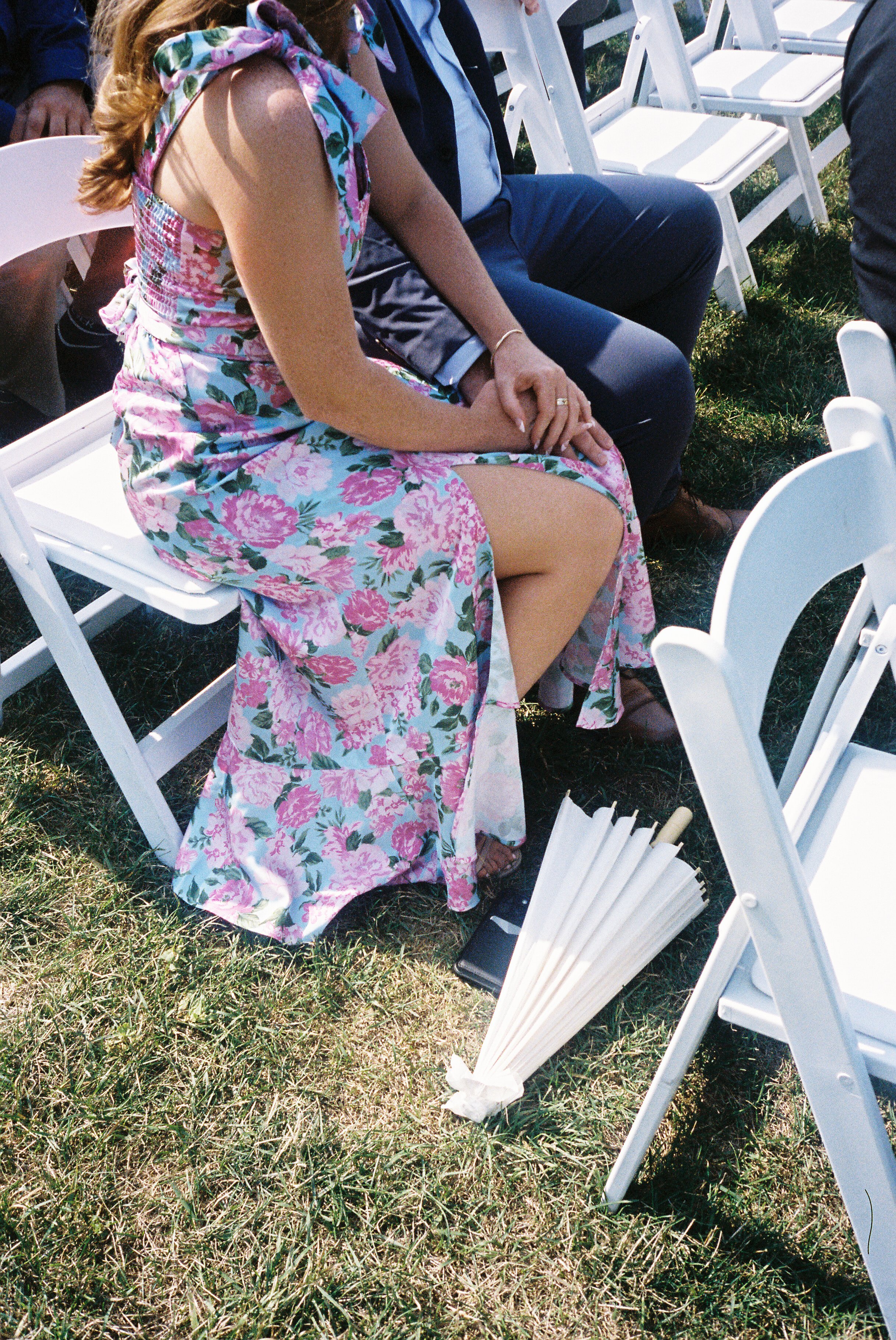 Wedding guest in a floral dress seated among white ceremony chairs during an outdoor vineyard wedding ceremony.