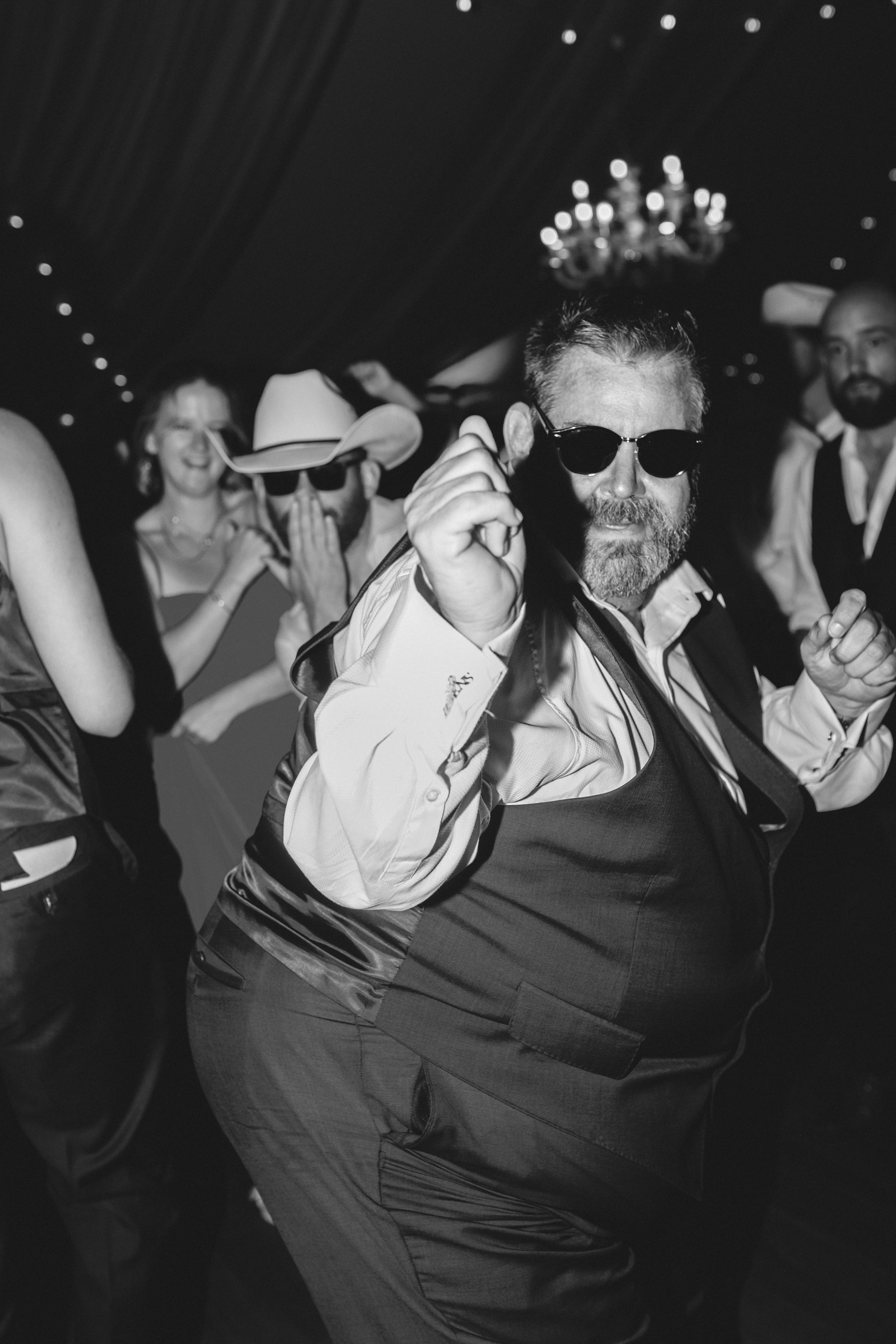 Black-and-white portrait of a wedding guest dancing on the reception dance floor beneath chandeliers and draped fabric.