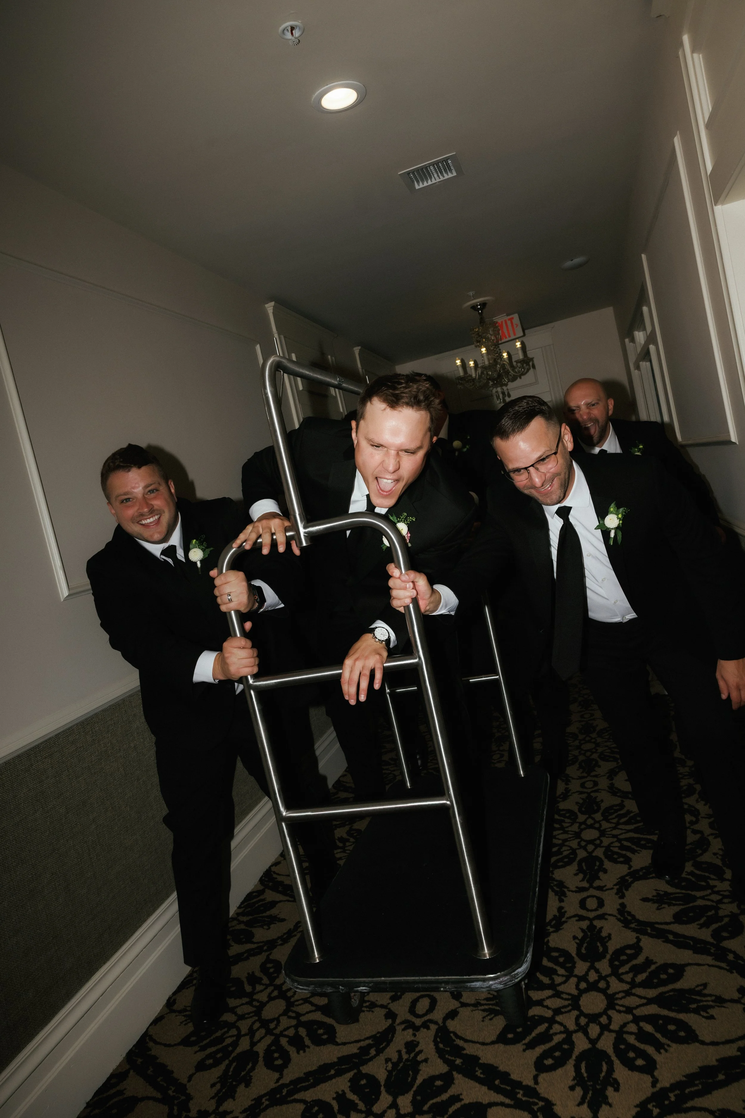 Groomsmen racing a luggage cart through the hotel hallway during a fun wedding day moment at Stafford's Perry Hotel.