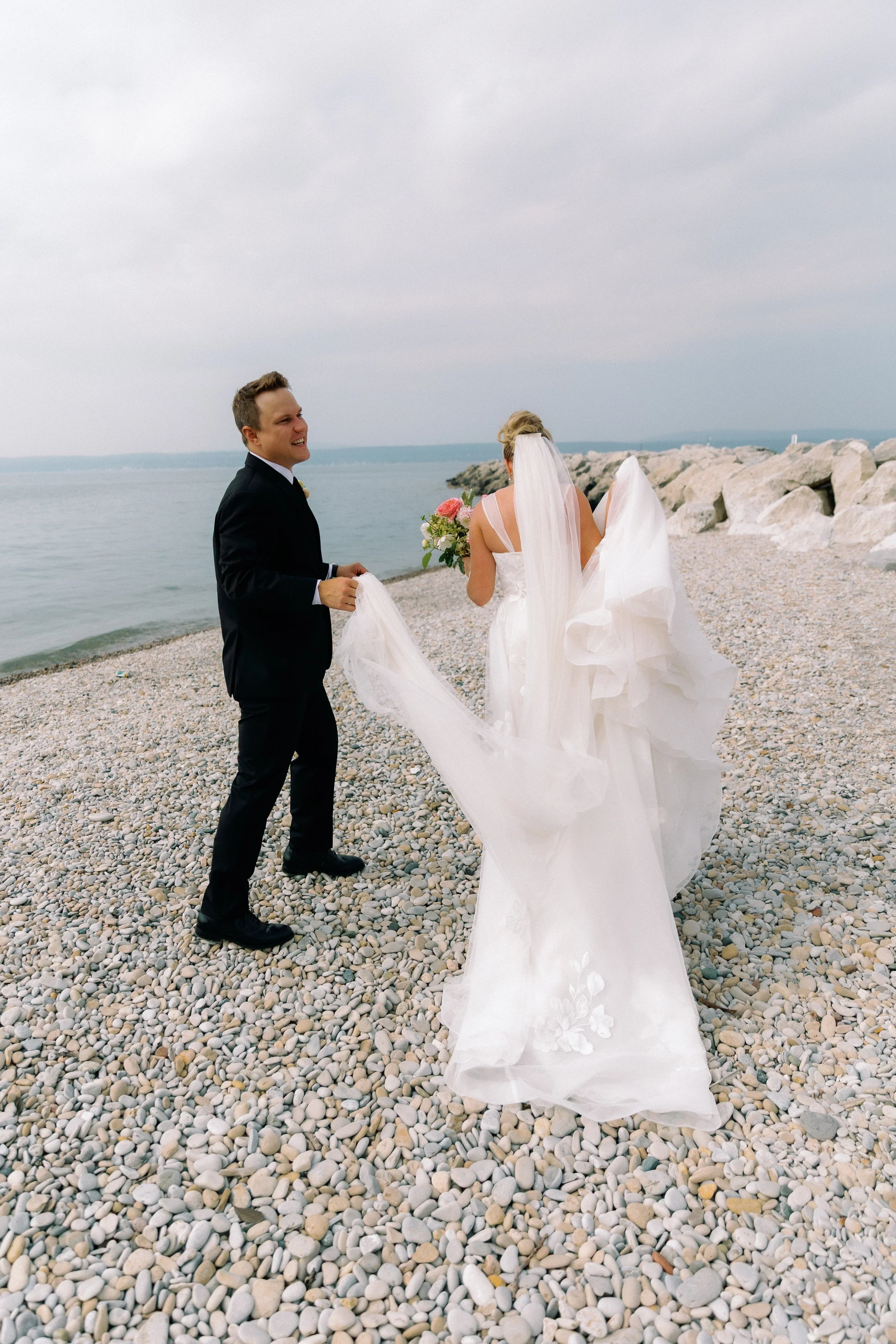 Newlyweds walking along the rocky shoreline near Stafford's Perry Hotel with the bride's veil flowing behind her.