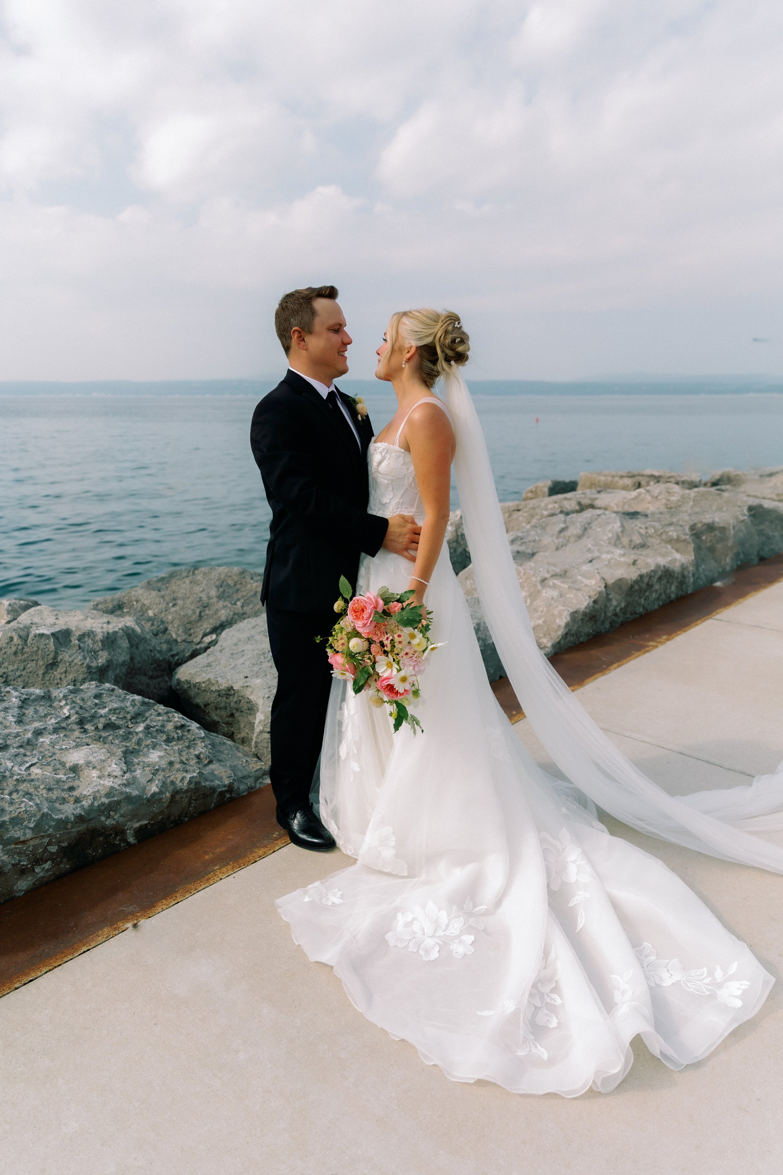 Bride and groom embracing by the waterfront with a colorful wedding bouquet overlooking Lake Michigan.