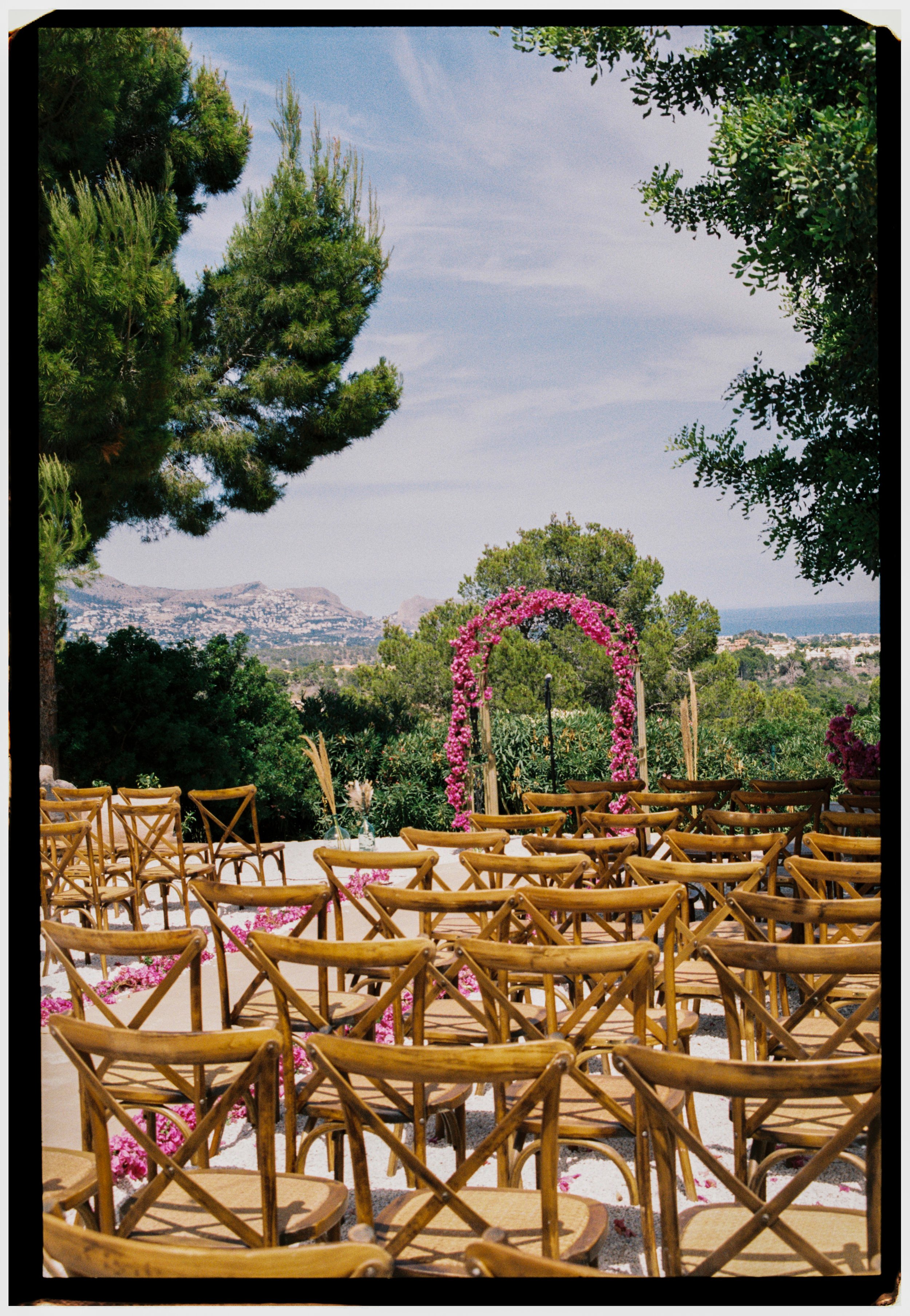 Outdoor ceremony setup with wooden chairs and a vibrant floral arch overlooking the Spanish countryside.