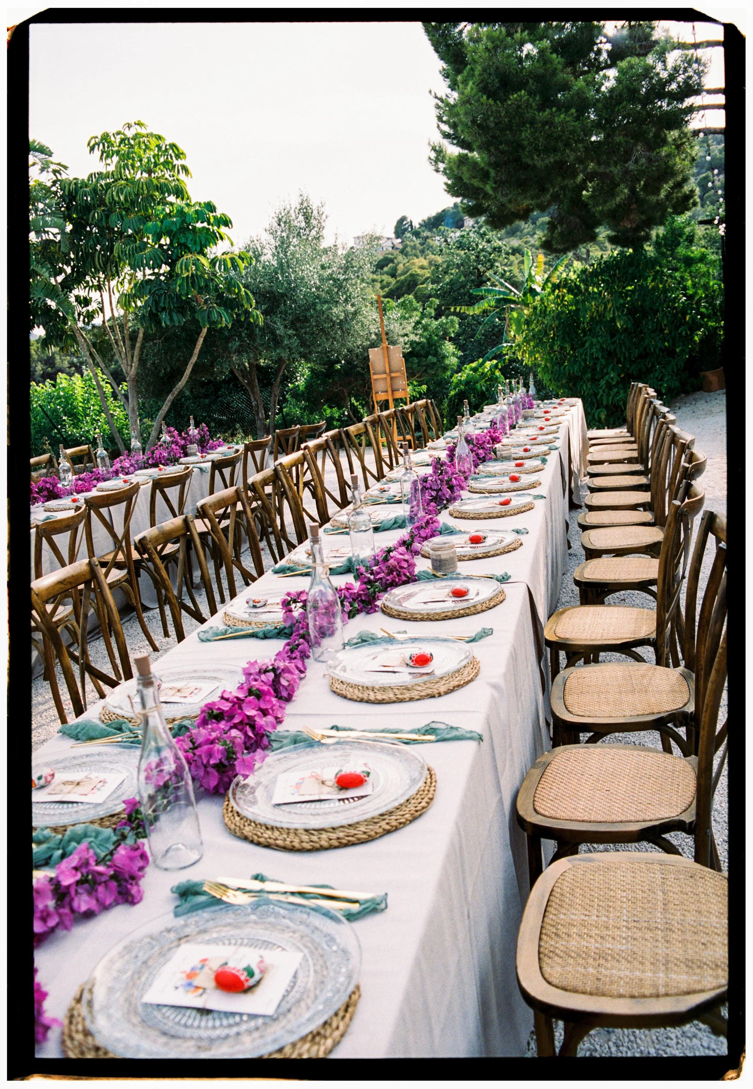Long outdoor reception table styled with purple florals, woven chargers, and elegant place settings.