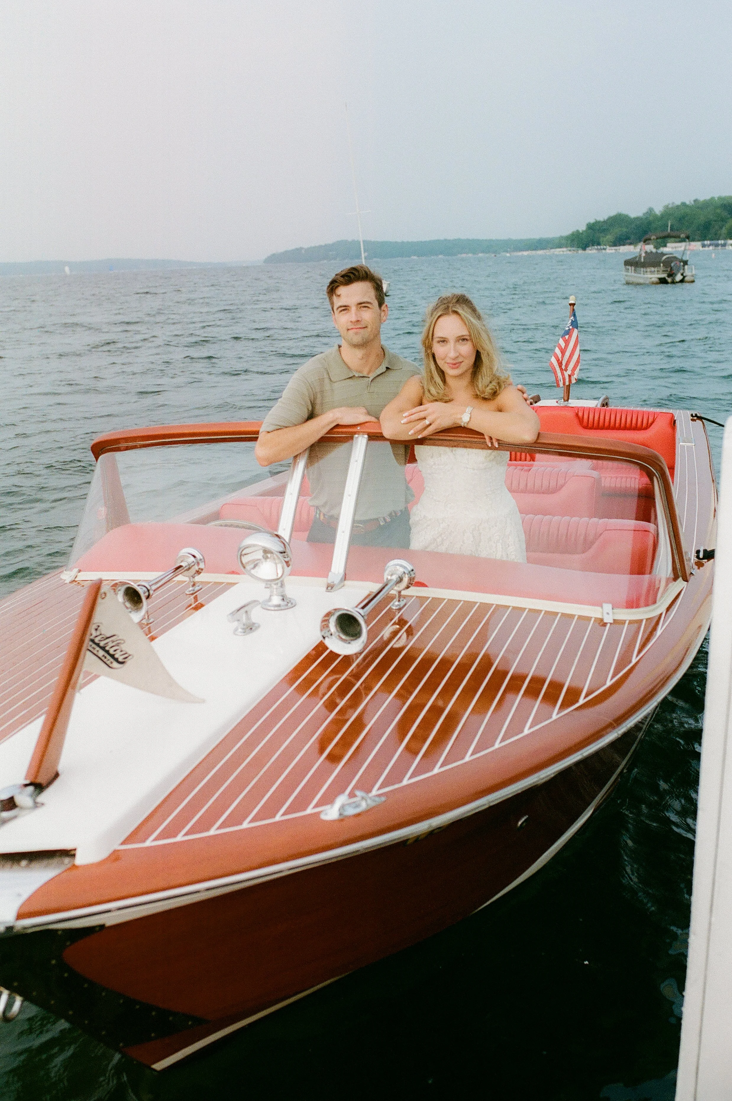 Couple posing at the front of a vintage wooden boat during their Lake Geneva Engagement session with calm water in the background.