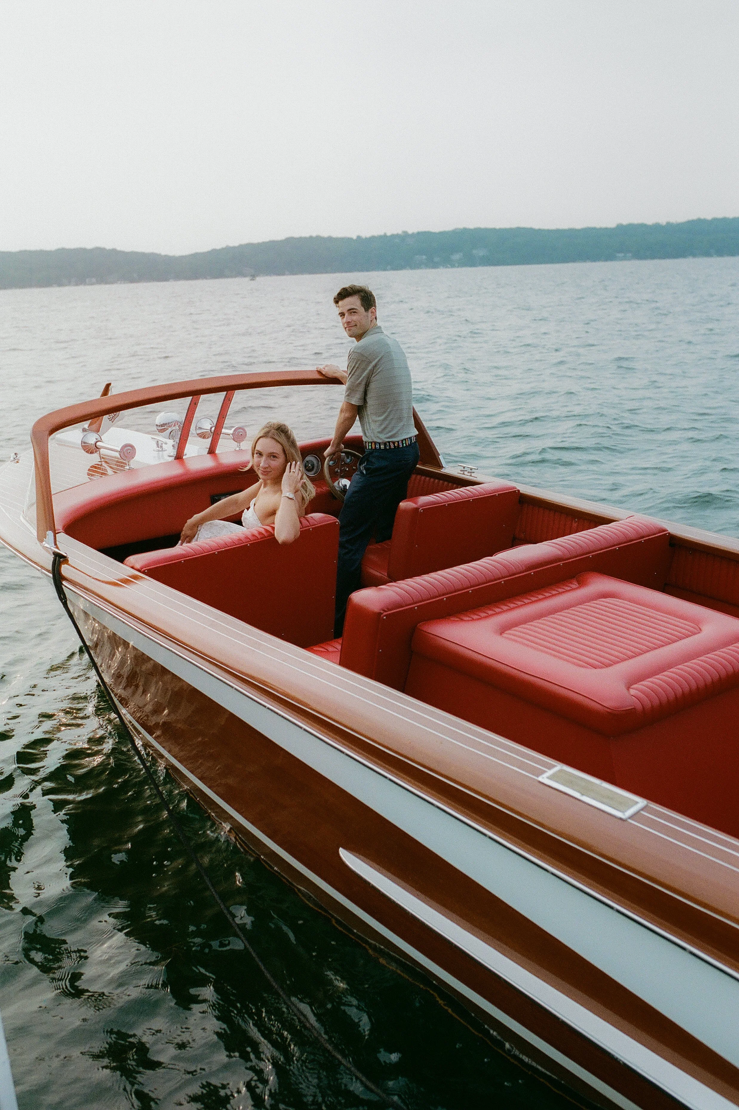 Bride and groom aboard a vintage wooden speedboat, smiling back toward the camera as they cruise across the water during sunset.