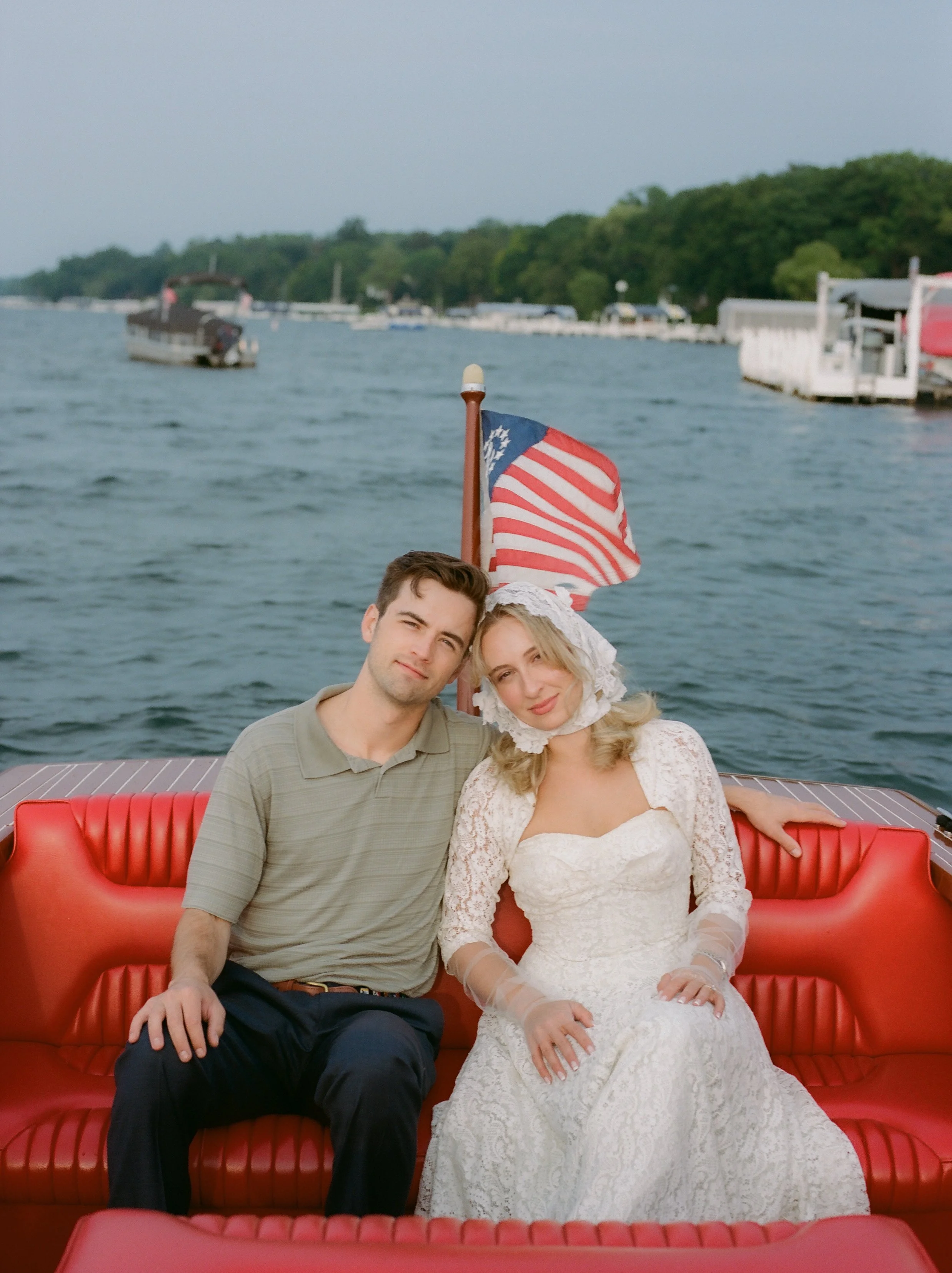 Engaged couple sitting together on a classic wooden boat with an American flag waving behind them on the lake.