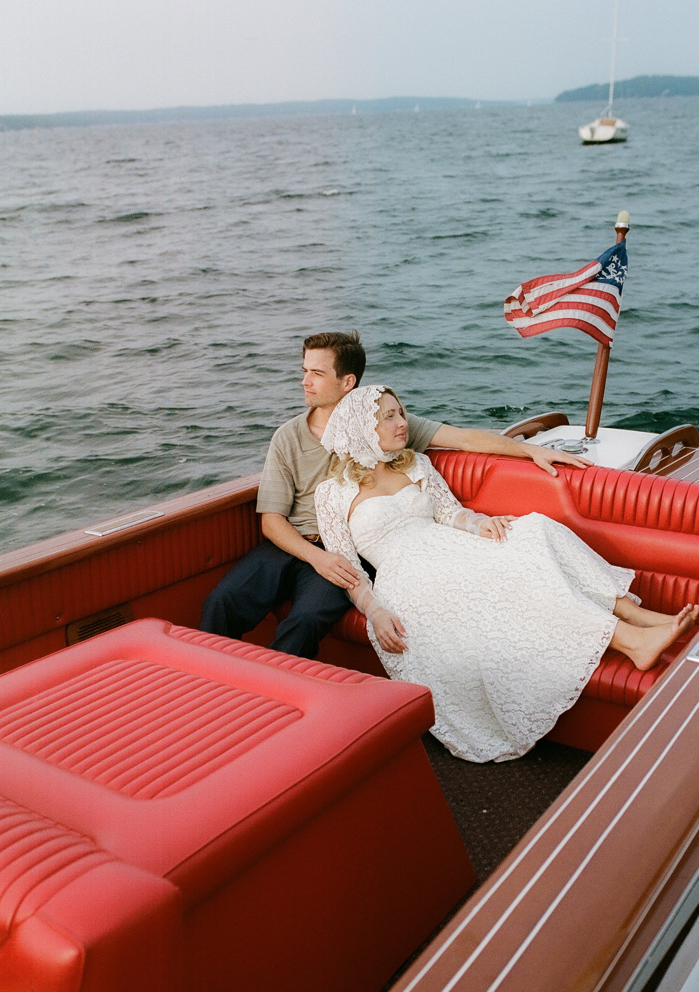 Bride and groom relaxing together on the red leather seating of a vintage wooden boat during a romantic lakeside engagement session.
