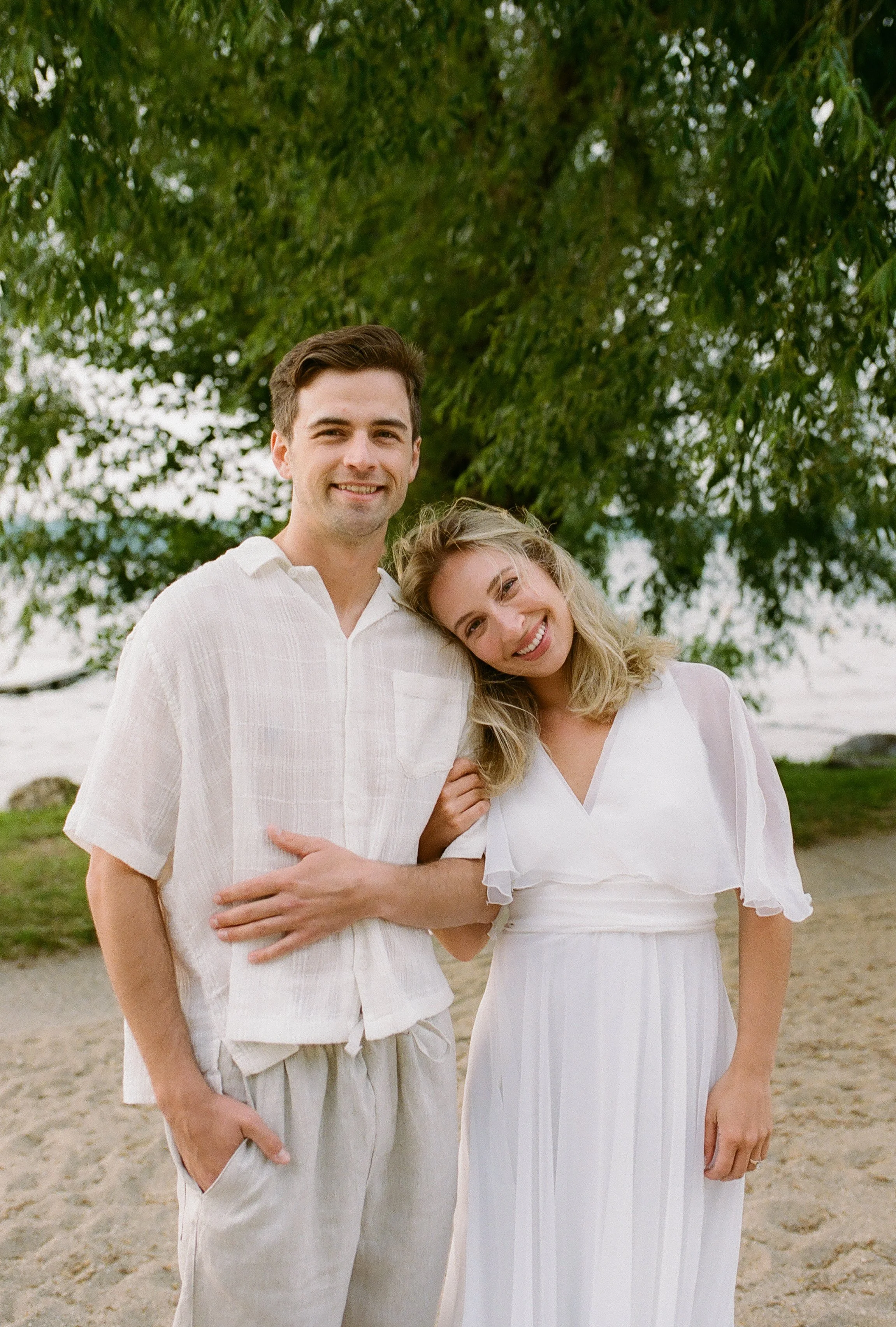 Engaged couple smiling together on the beach in light, neutral outfits beneath a leafy tree.