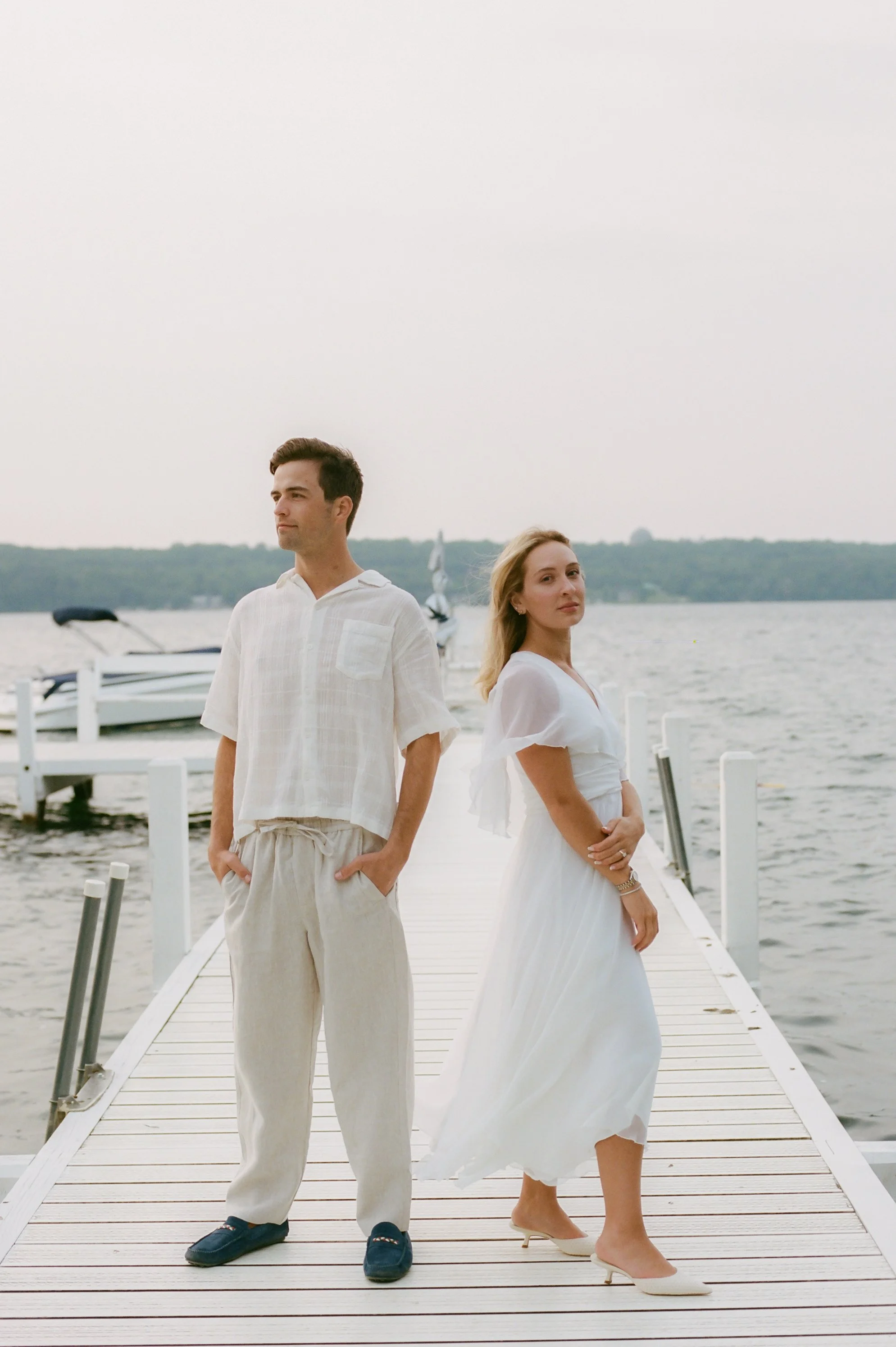 Engaged couple standing back-to-back on a lakeside dock during their Lake Geneva Engagement session at sunset.