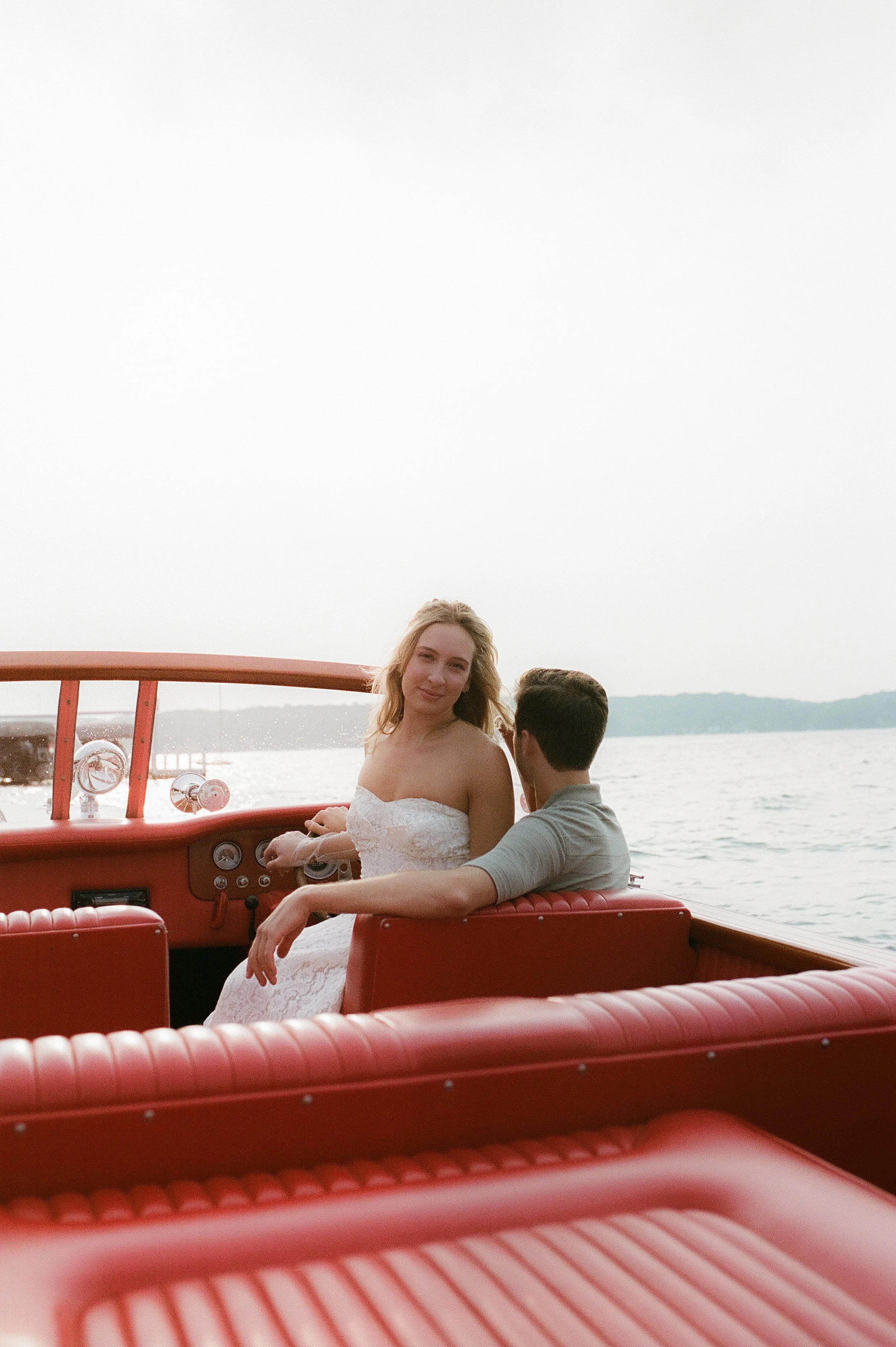 Bride smiling toward the camera while sitting aboard a vintage wooden boat with her fiancé on the lake at sunset.