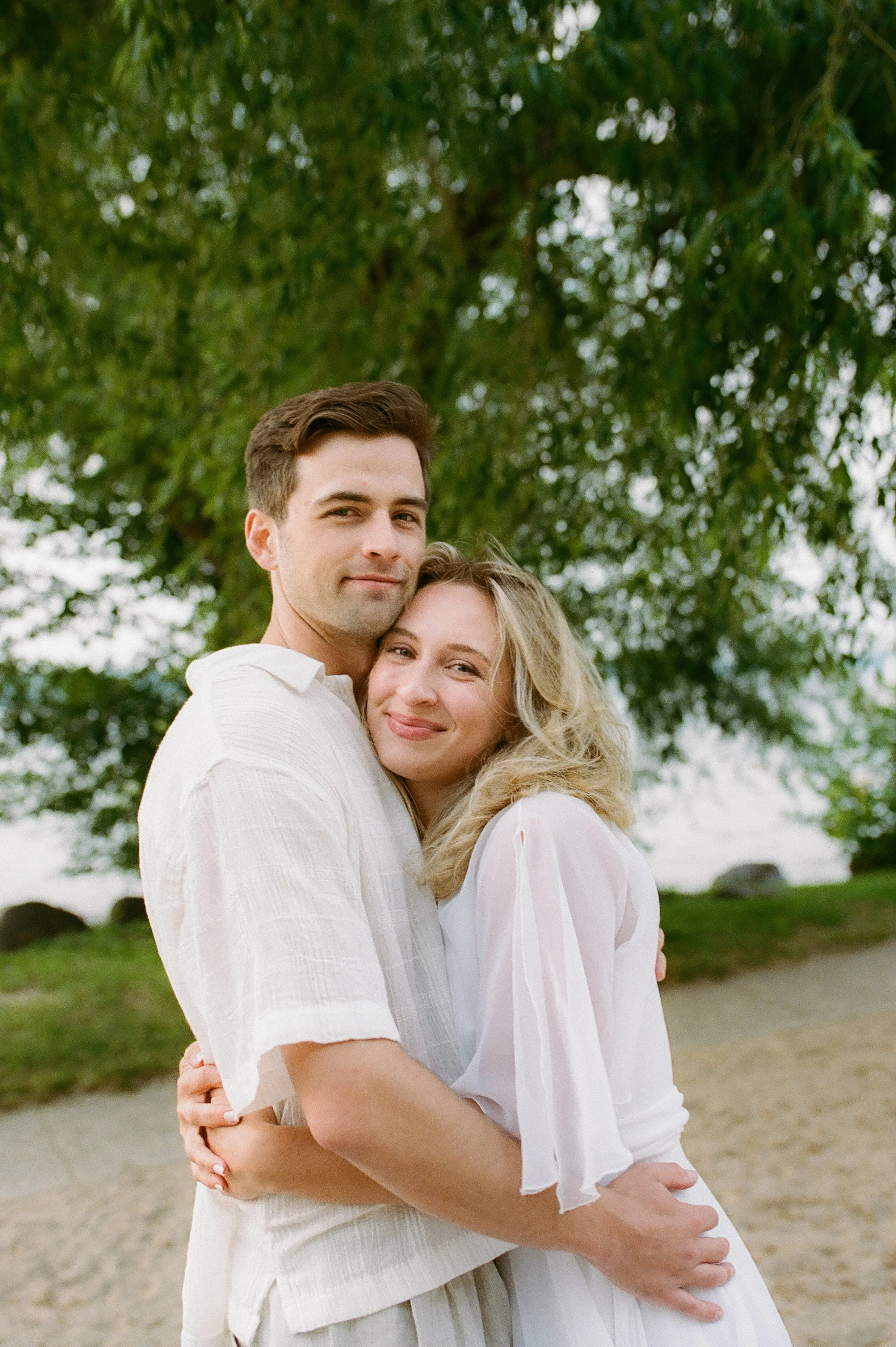 Engaged couple embracing beneath a leafy tree with soft smiles during their Lake Geneva Engagement session.