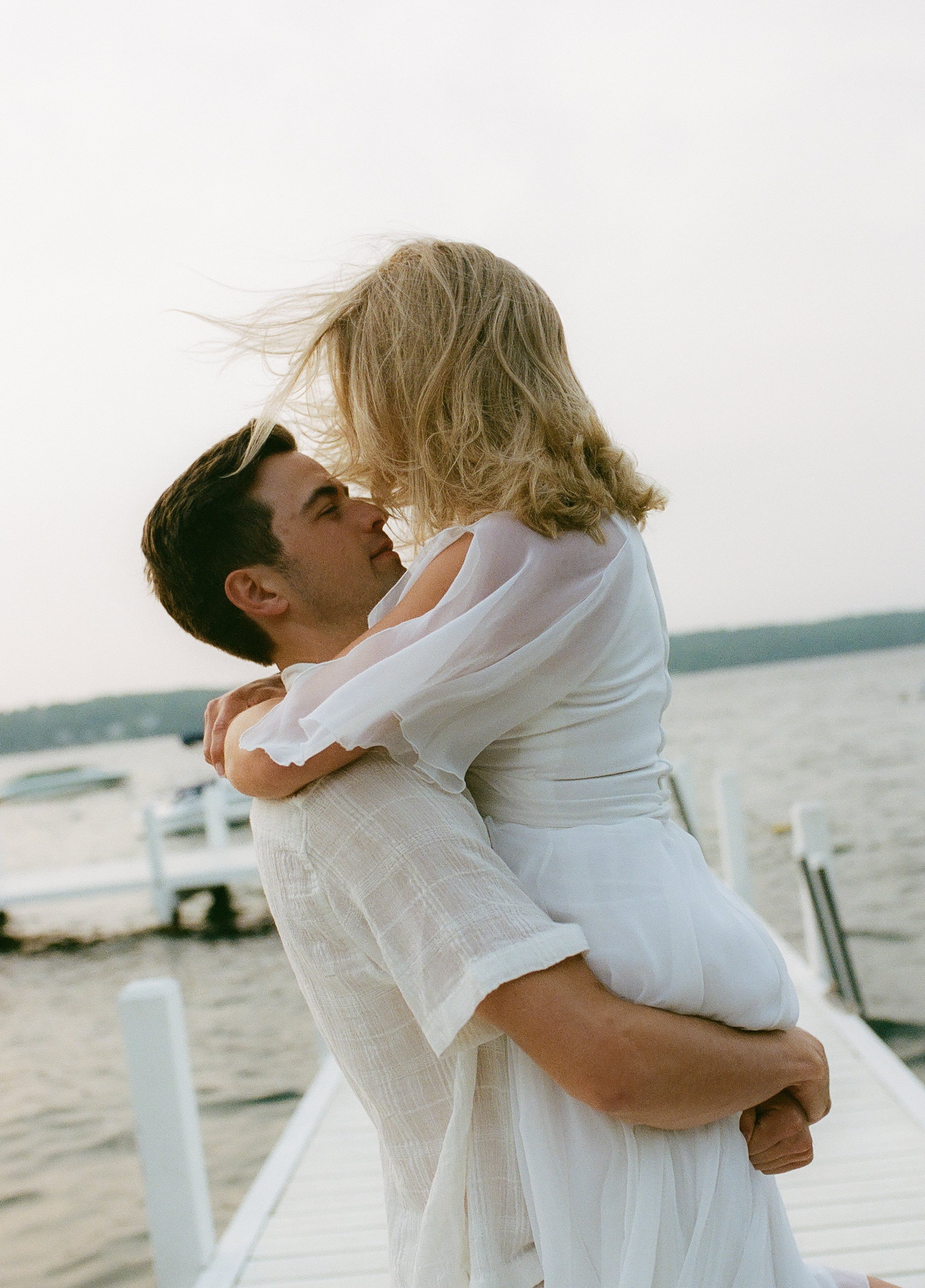 Groom lifting his fiancée into an embrace on a lakeside dock at golden hour.