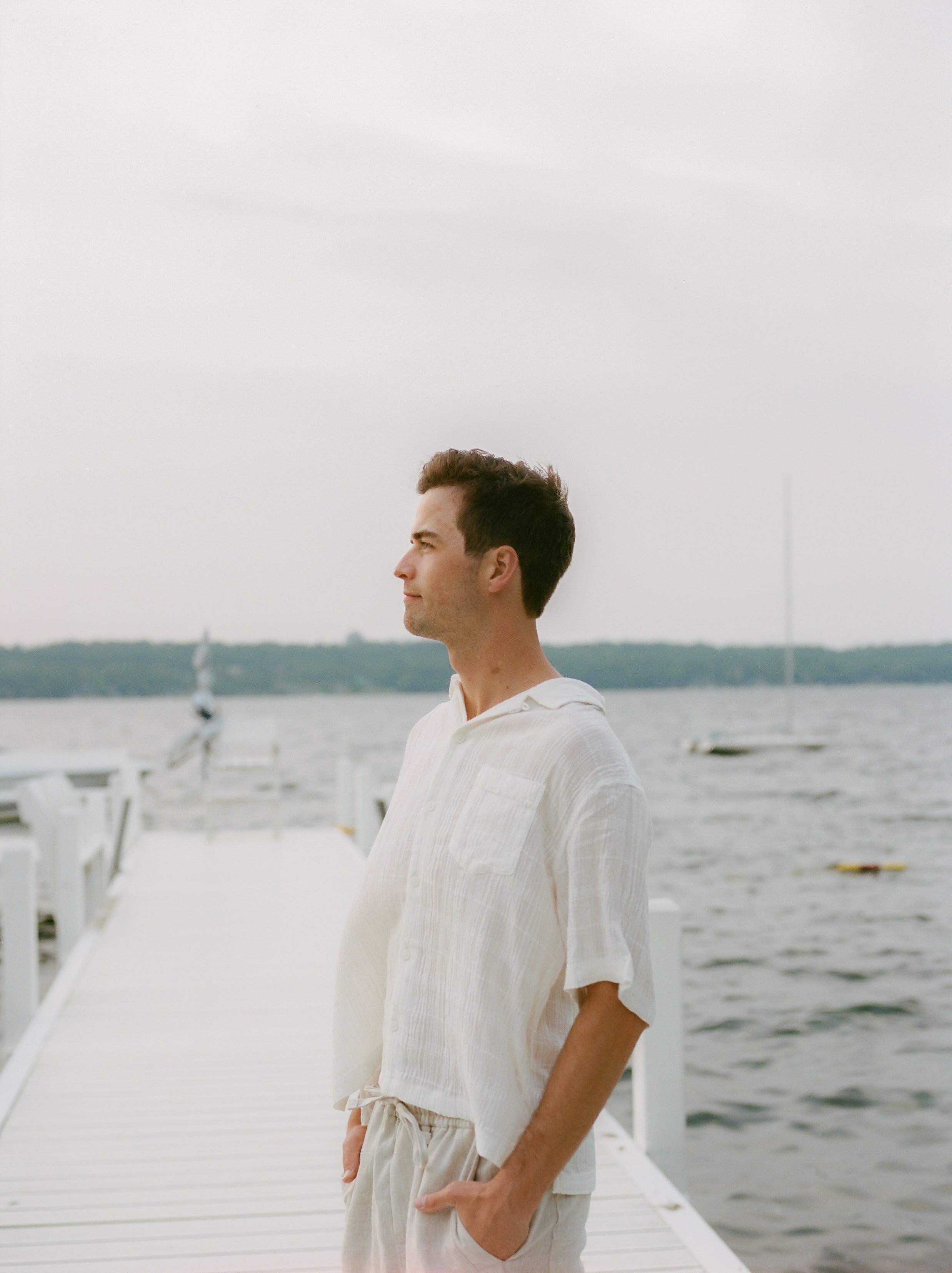 Groom standing on a lakeside dock overlooking Lake Geneva during sunset in a relaxed engagement portrait.
