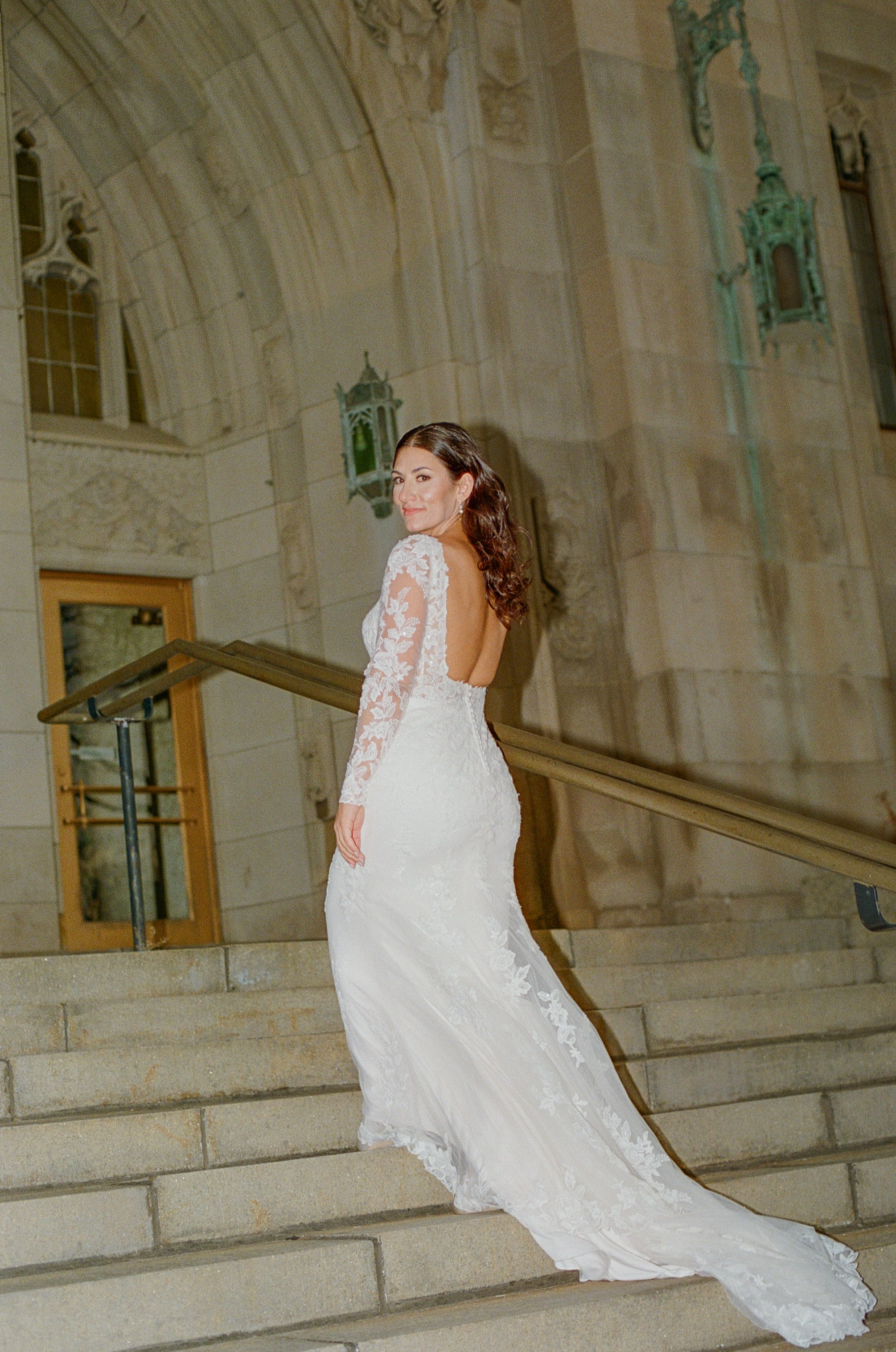 Bride showing the open-back lace detail of her long-sleeve wedding dress on stone steps.