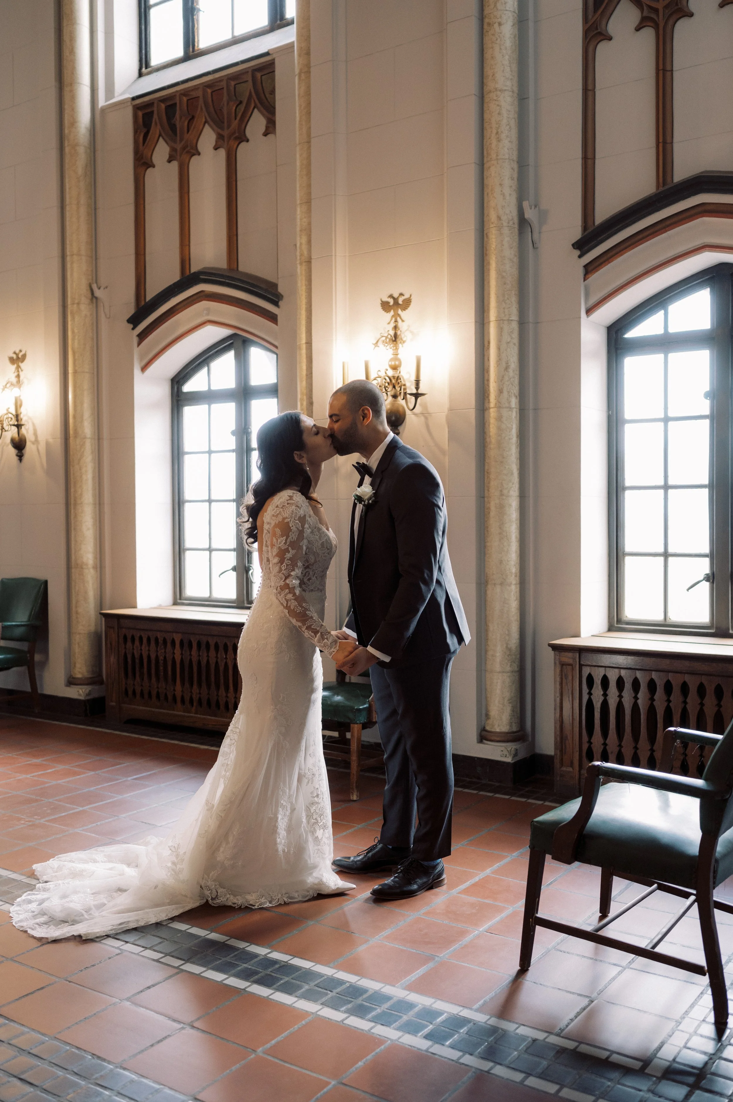 Newlyweds smiling and holding hands beside a large window during their downtown Detroit wedding.