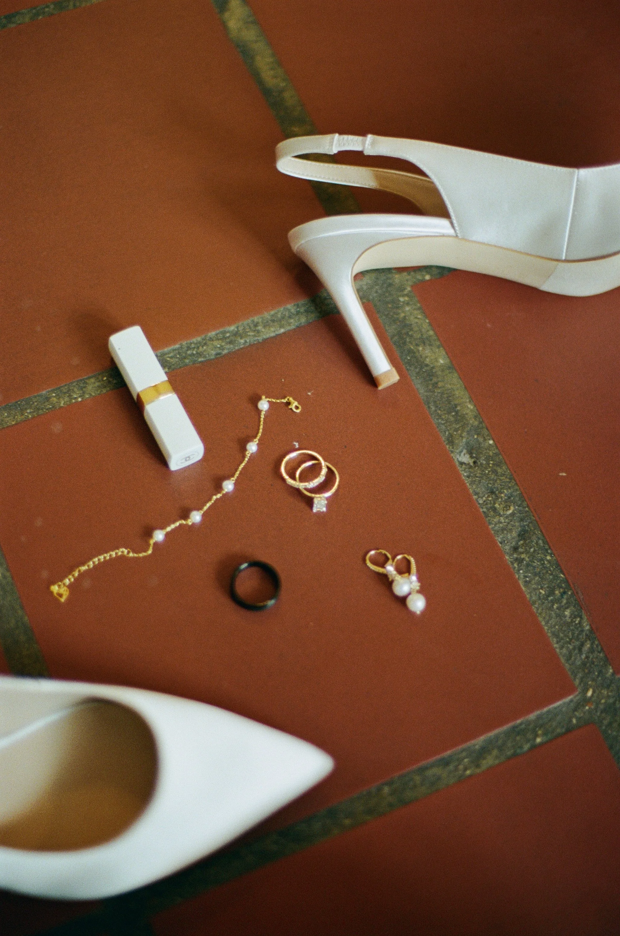 Bridal details featuring white heels, wedding rings, jewelry, and perfume arranged on a tiled floor.
