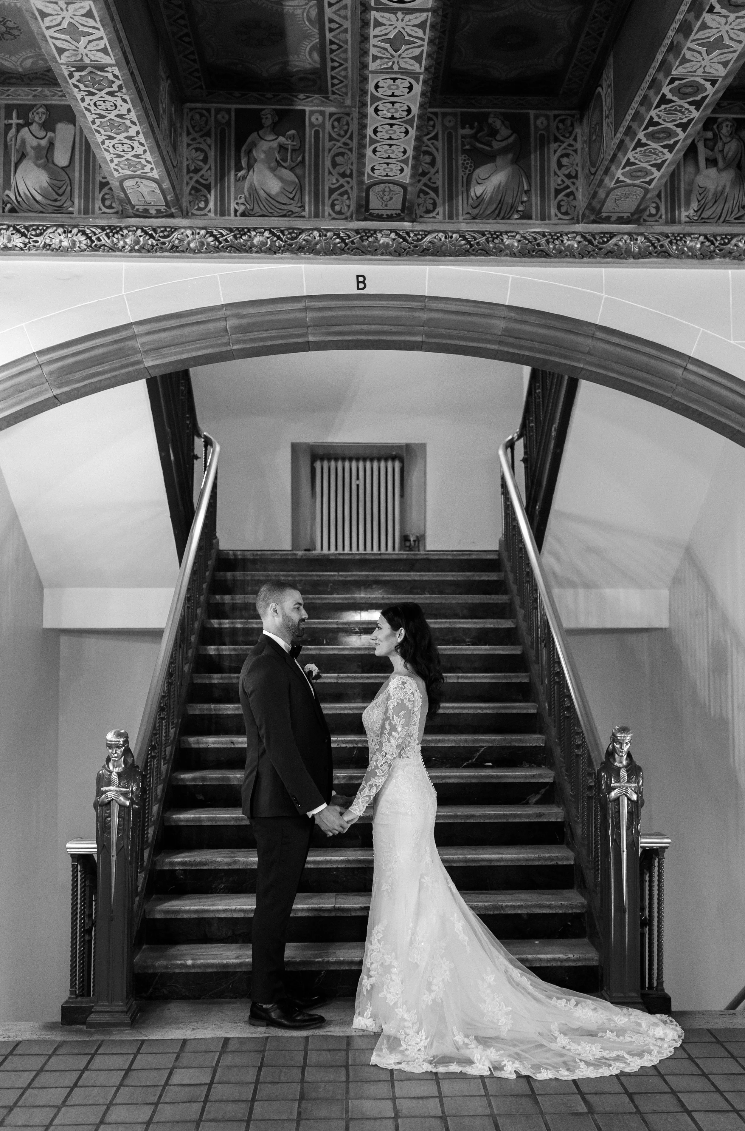 Black-and-white wedding portrait of the bride and groom holding hands in front of a grand historic staircase.