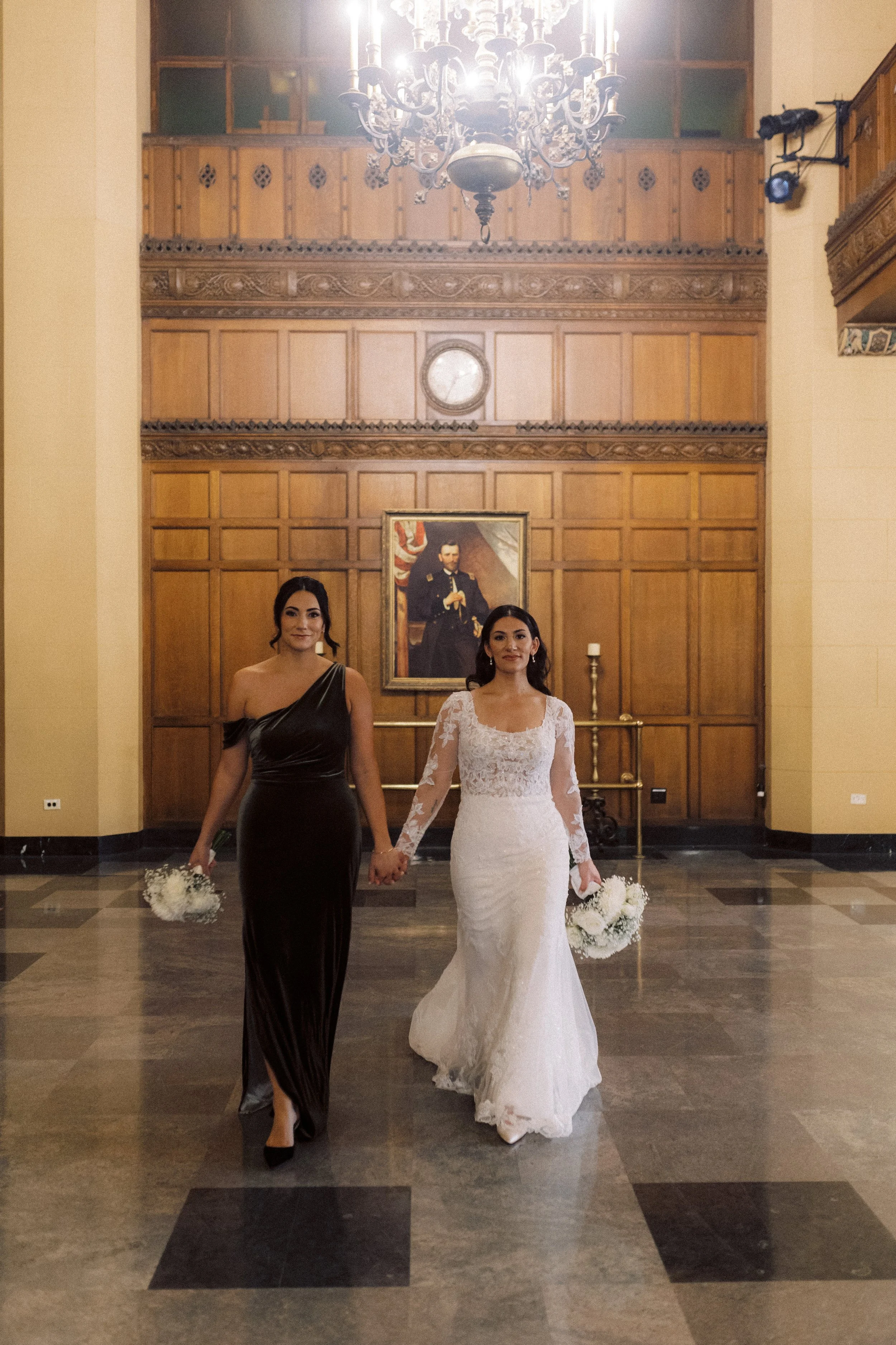 Bride walking hand in hand with her bridesmaid while carrying a white floral wedding bouquet.