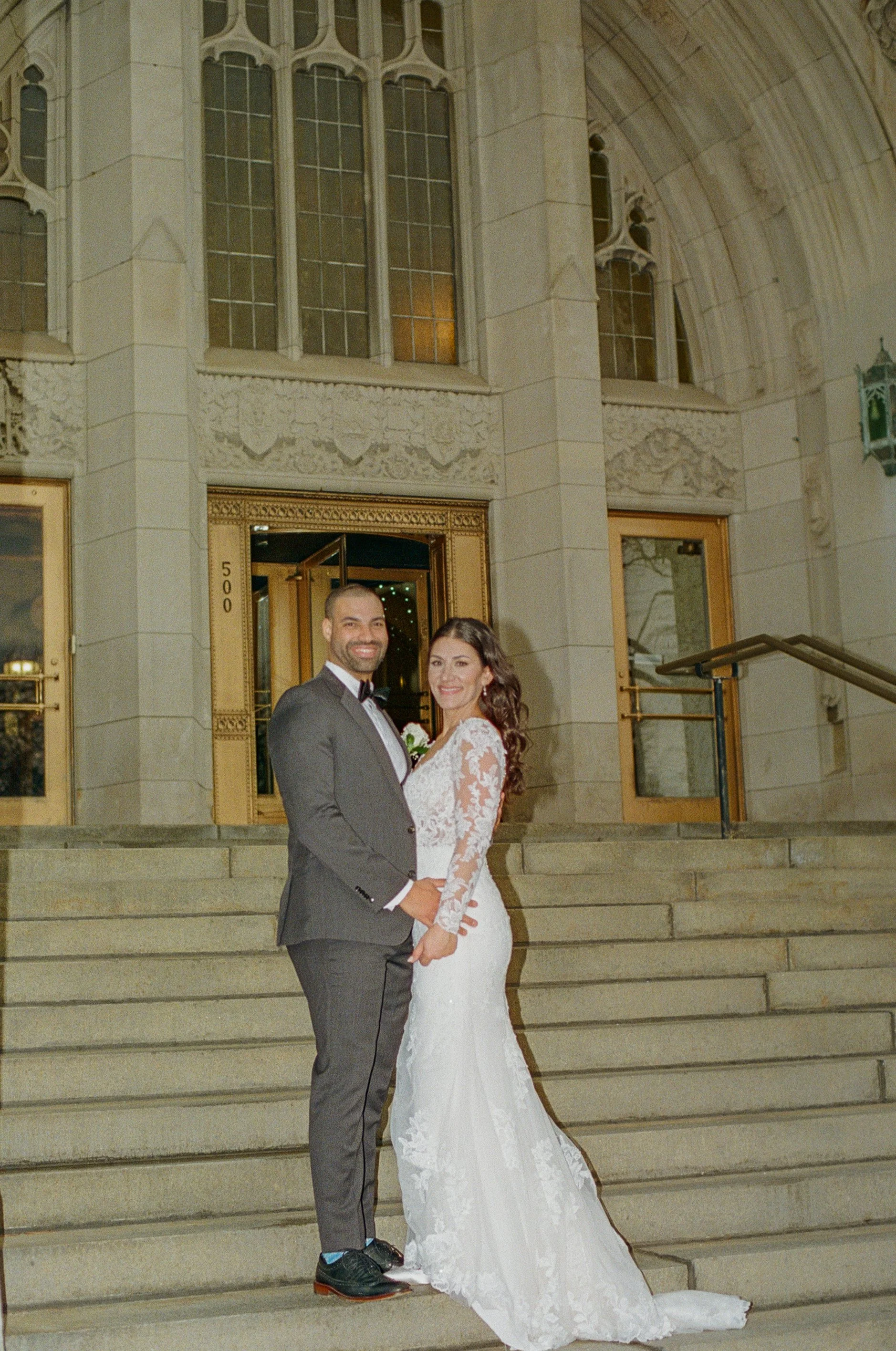 A bride and groom smile at the camera on the steps of The Masonic Temple in downtown Detroit