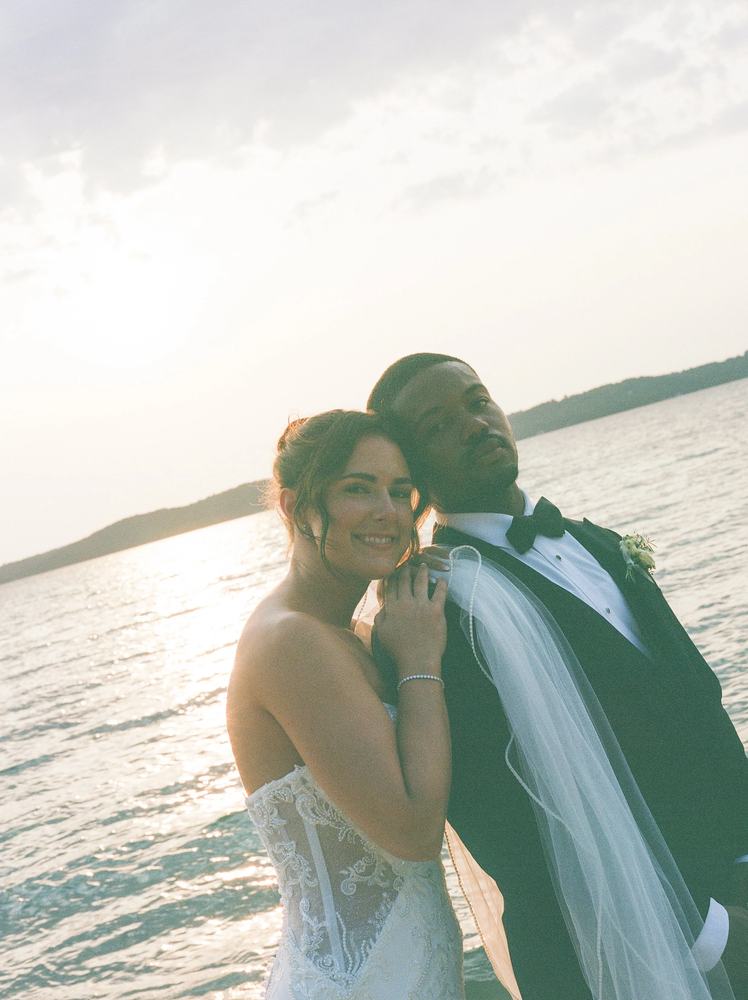 Newlyweds smiling together by the water with the bride’s veil draped over the groom’s shoulder.