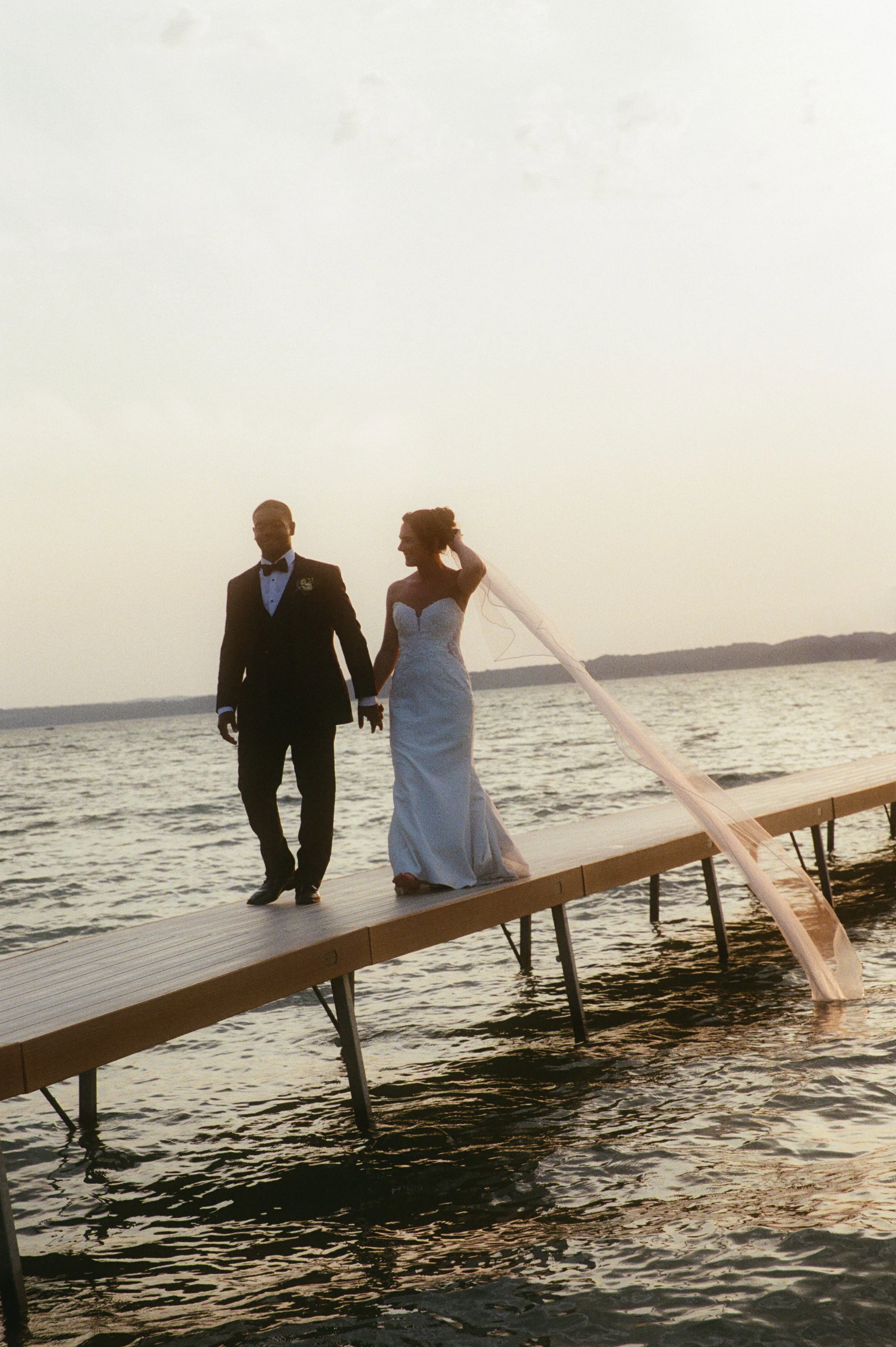 Bride and groom walking hand in hand along a dock over the lake at sunset.