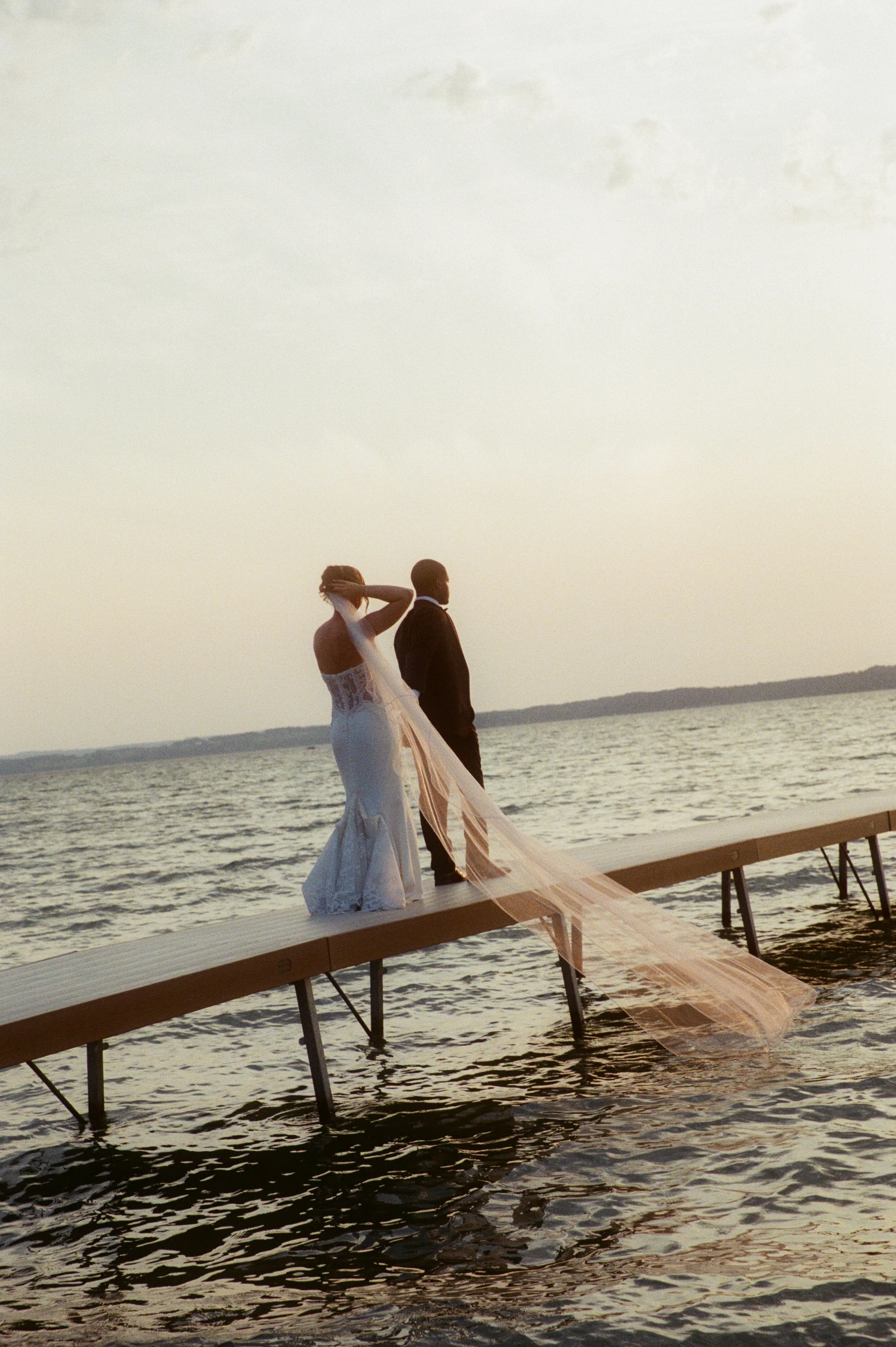 Bride and groom standing together on a dock overlooking the lake at sunset.
