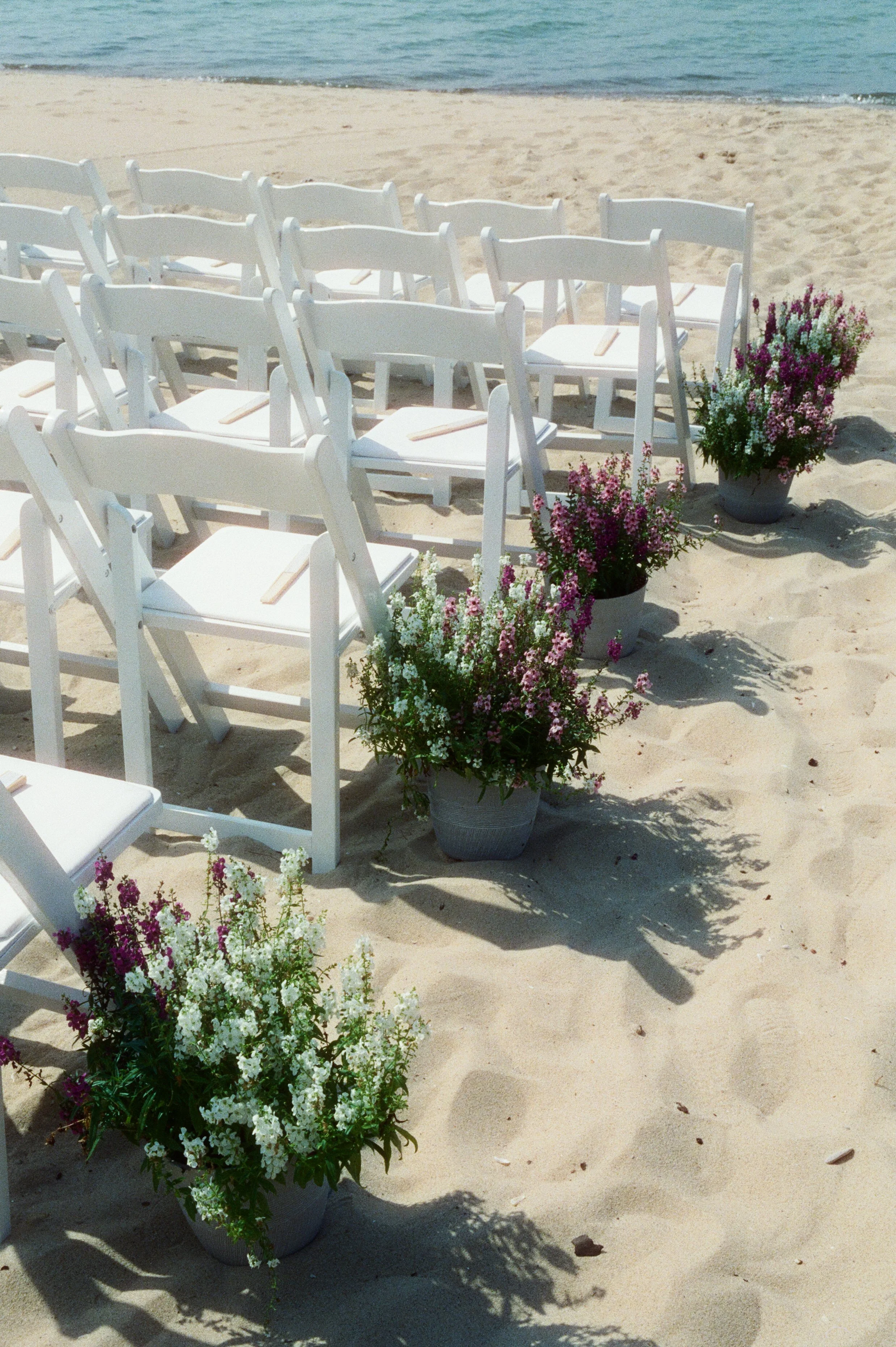 White ceremony chairs lined with colorful potted flowers along the sandy lakeside wedding aisle.
