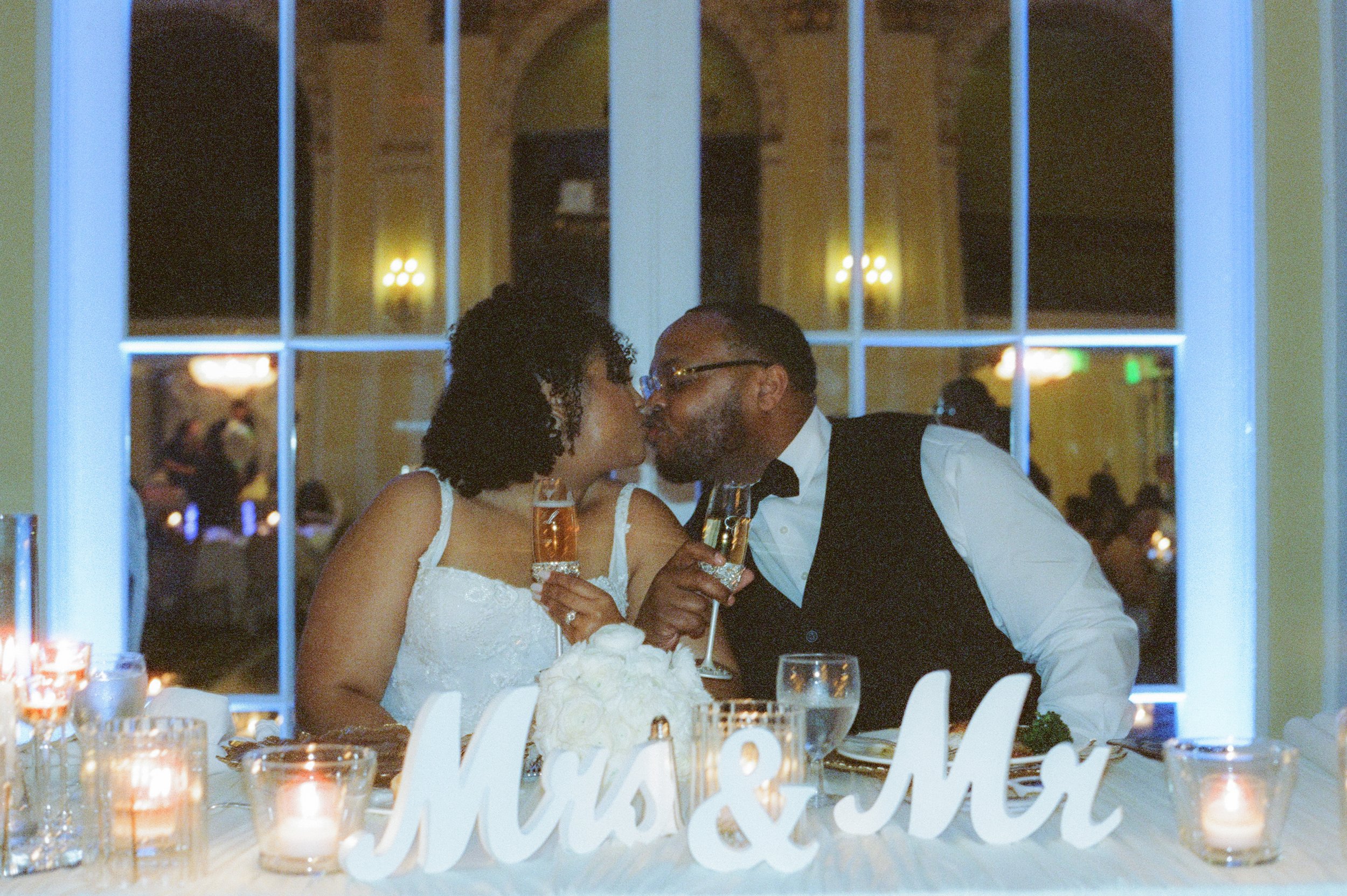 Bride and groom sharing a kiss while holding champagne glasses at their sweetheart table captured in cinematic film photography style