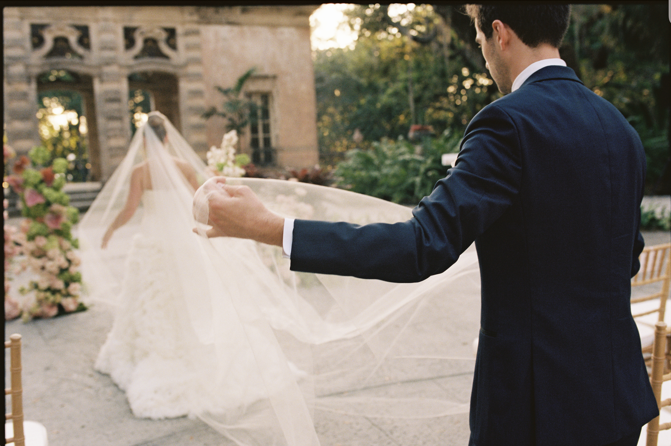 Groom lifting the bride’s veil as she walks toward the ceremony space during their Vizcaya Museum and Gardens wedding.