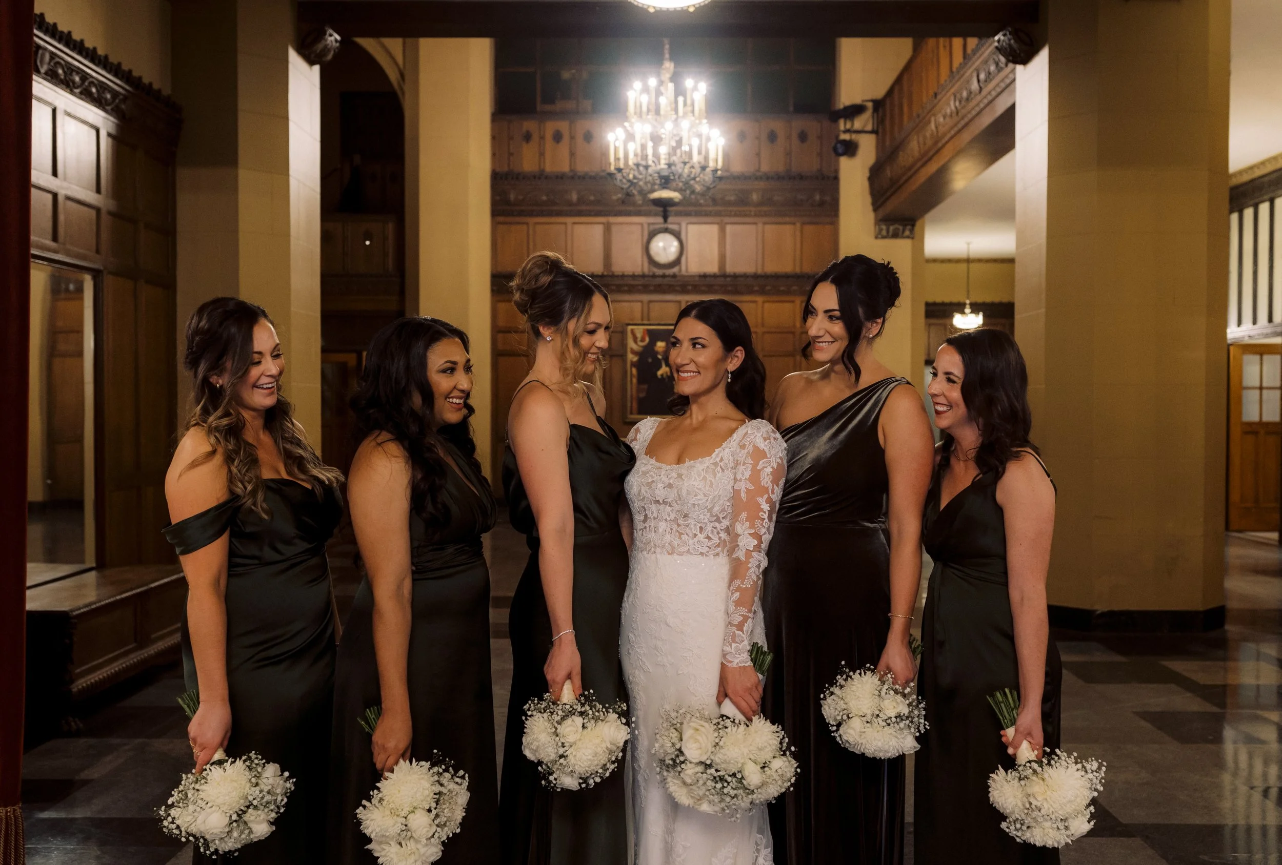 Bride smiling with her bridesmaids in dark dresses and white bouquets before her downtown Detroit wedding ceremony.