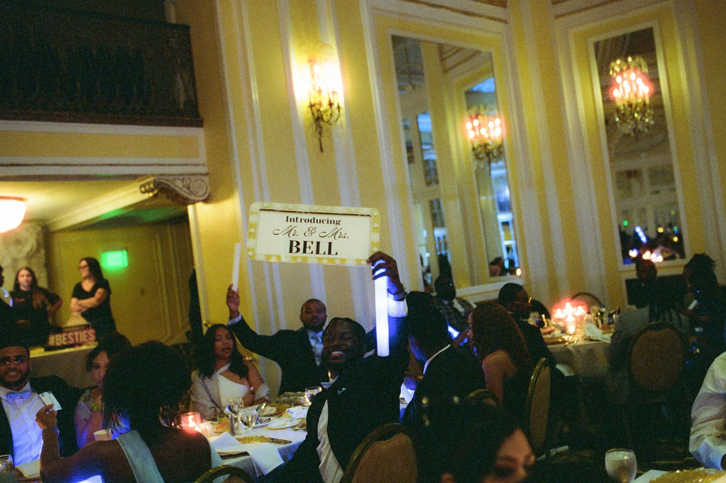 Wedding guests celebrating at the reception while holding glow sticks and a sign introducing Mr. and Mrs. Bell